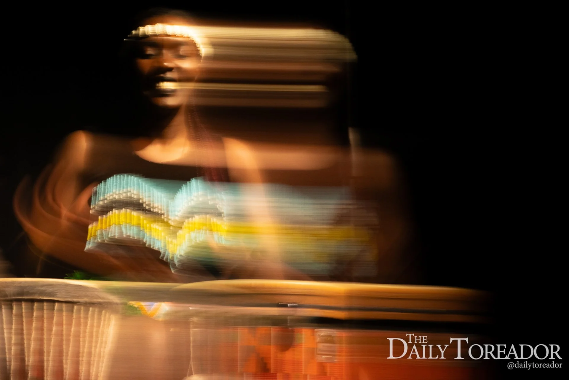 Blurred image of a person with dark skin and short hair, standing in front of dark background, with streaks of light and color creating an abstract effect. The image includes the text "The Daily Toreador" and social media handle "@dailytoreador" at t