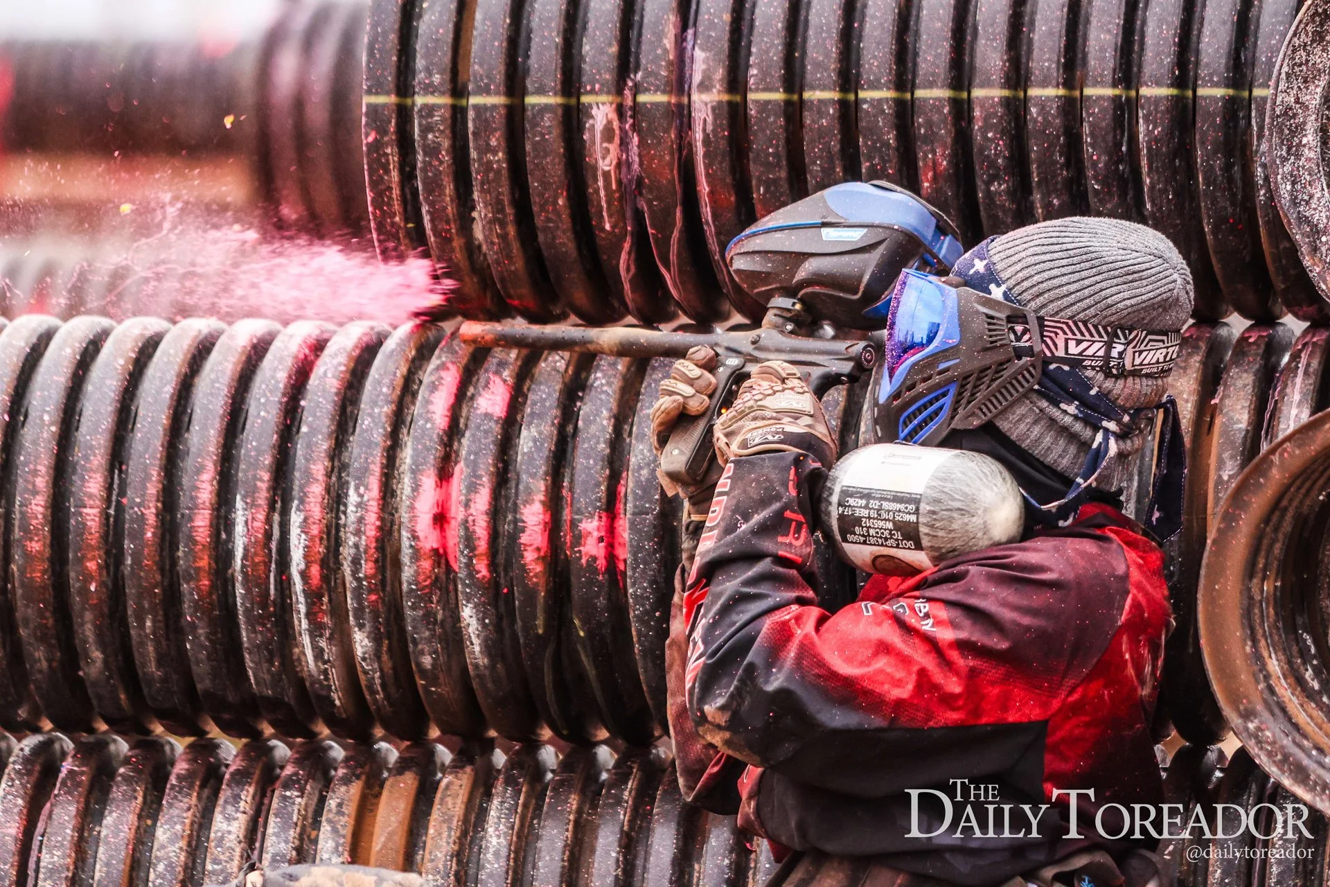 A worker in red and black protective gear, including gloves and a gray knit hat, is welding a large, rusted metal pipe with a helmet and blue face shield. Sparks and bright light emanate from the welding process.