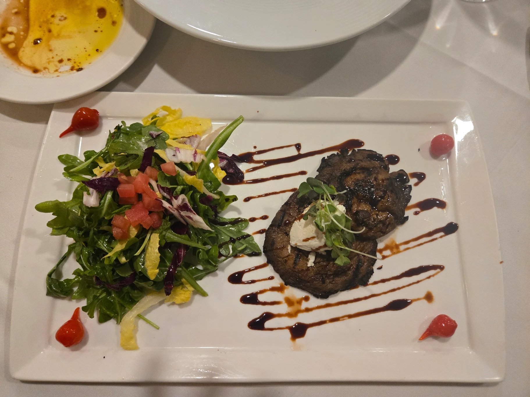 A white square plate with a grilled meat dish topped with microgreens, and a side salad of mixed greens, shredded yellow cheese, cherry tomatoes, and diced tomatoes, garnished with a drizzle of balsamic glaze. There are small dollops of ketchup aroun