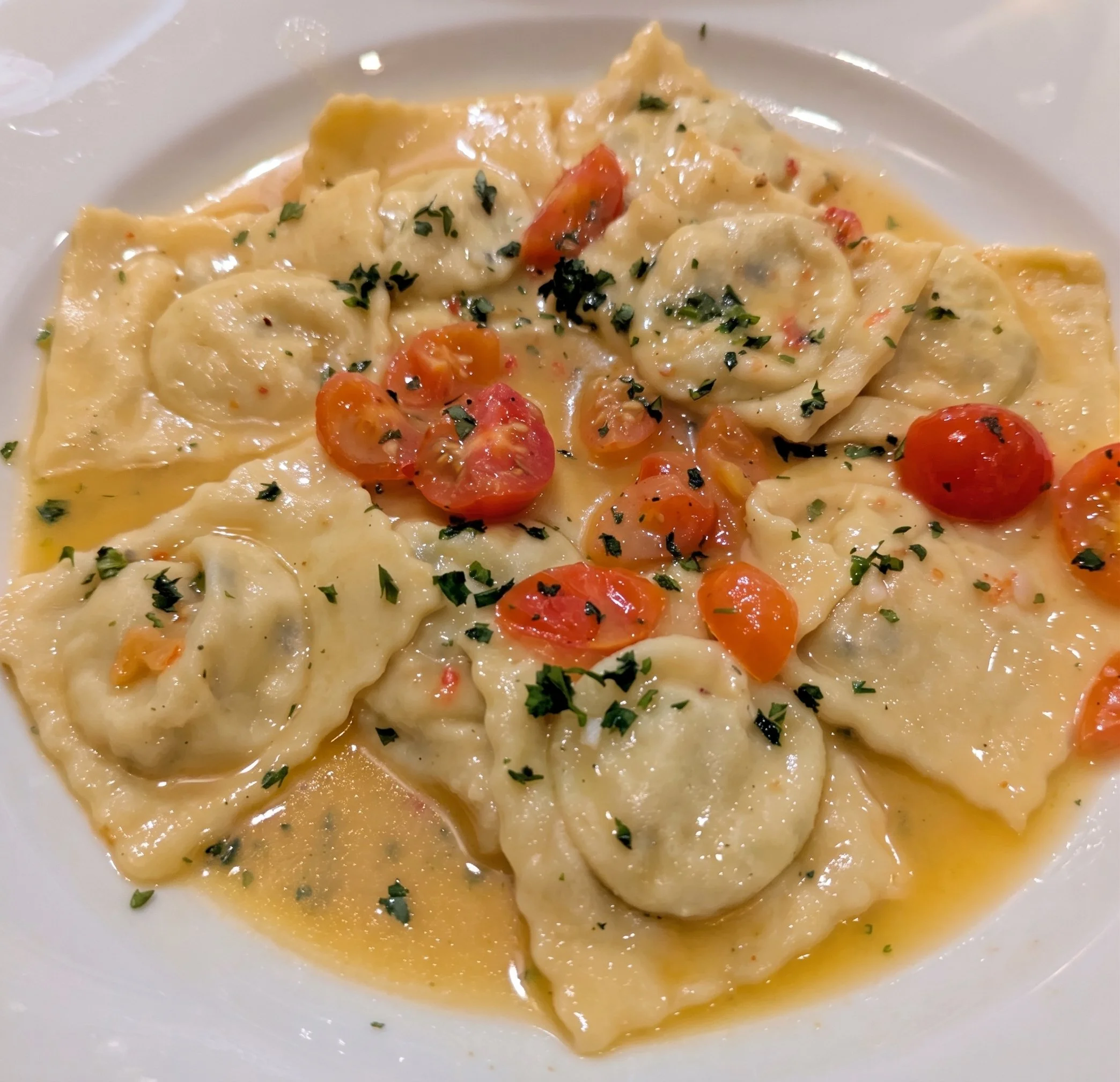 Close-up of a bowl of cheese ravioli pasta with cherry tomatoes, garnished with chopped herbs and drizzled with sauce.