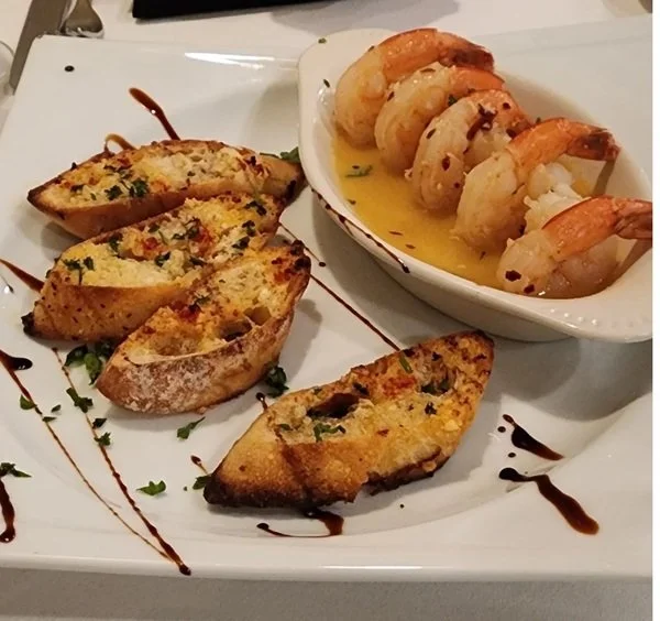 Appetizer plate with toasted garlic bread slices garnished with herbs and a drizzle of balsamic glaze, served alongside a small dish of cooked shrimp in butter sauce.