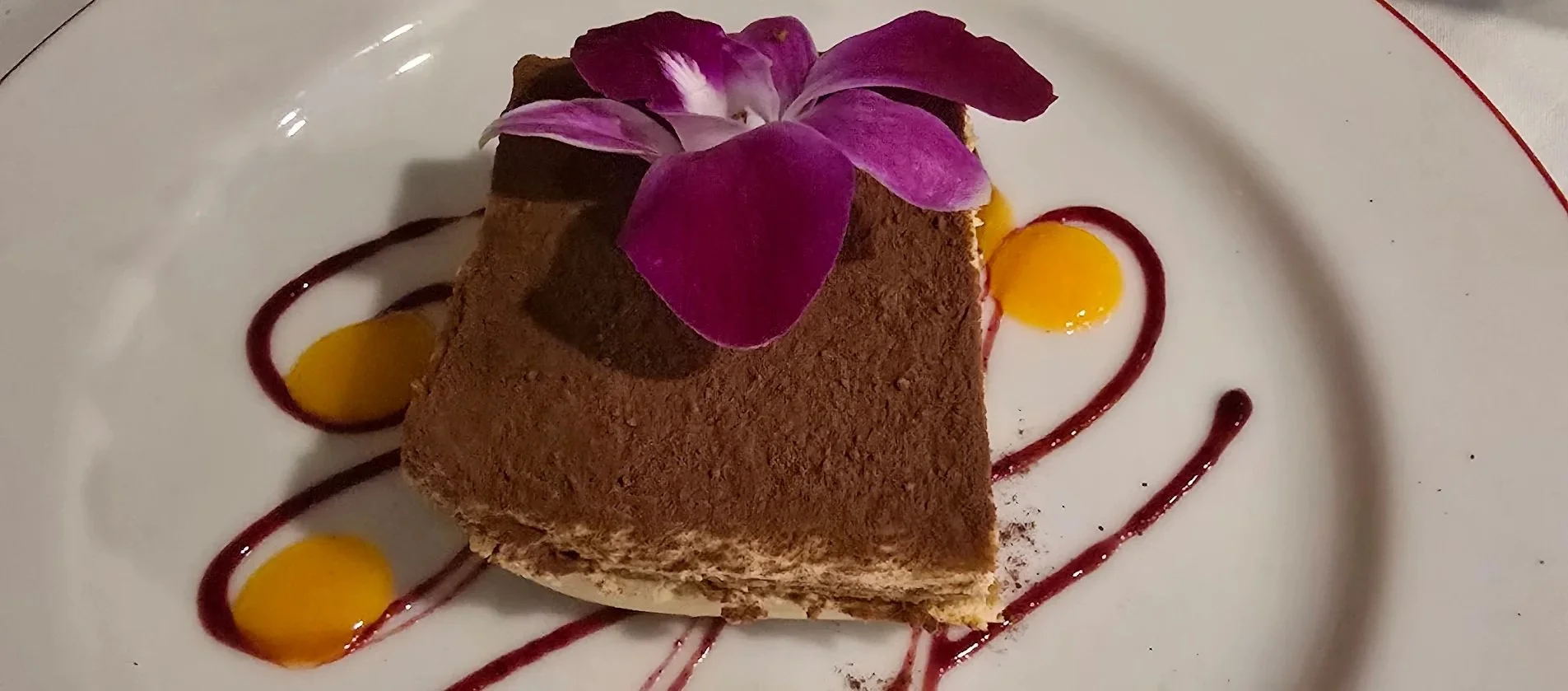 A slice of chocolate dessert garnished with a purple orchid flower, decorated with yellow and red sauce on a white plate.