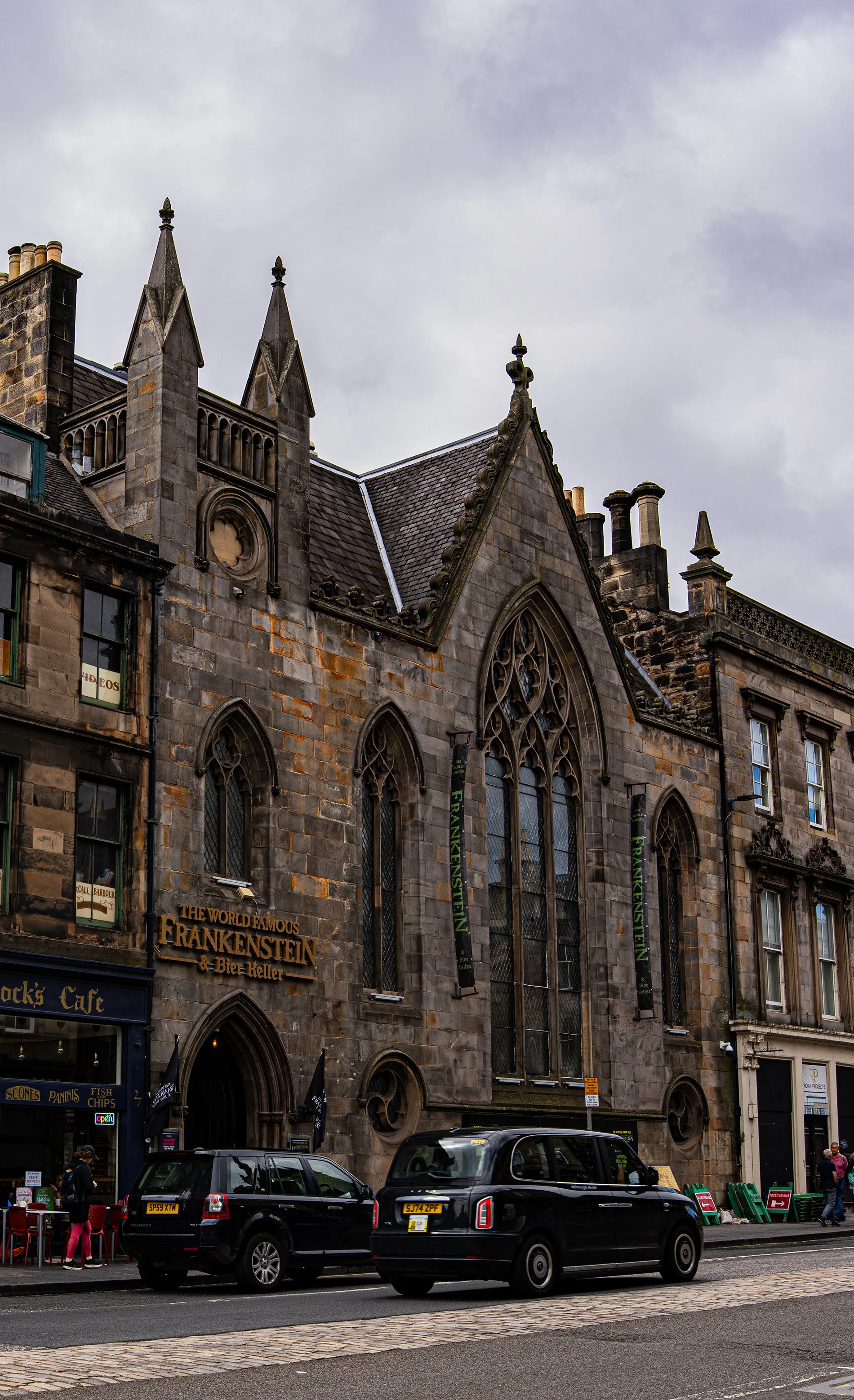 Fachada de un edificio gótico de piedra con ventanas altas y decorativas, y un cartel que dice "The World Famous Frankenstein & Bier Keller". En la calle hay autos estacionados y personas caminando afuera.