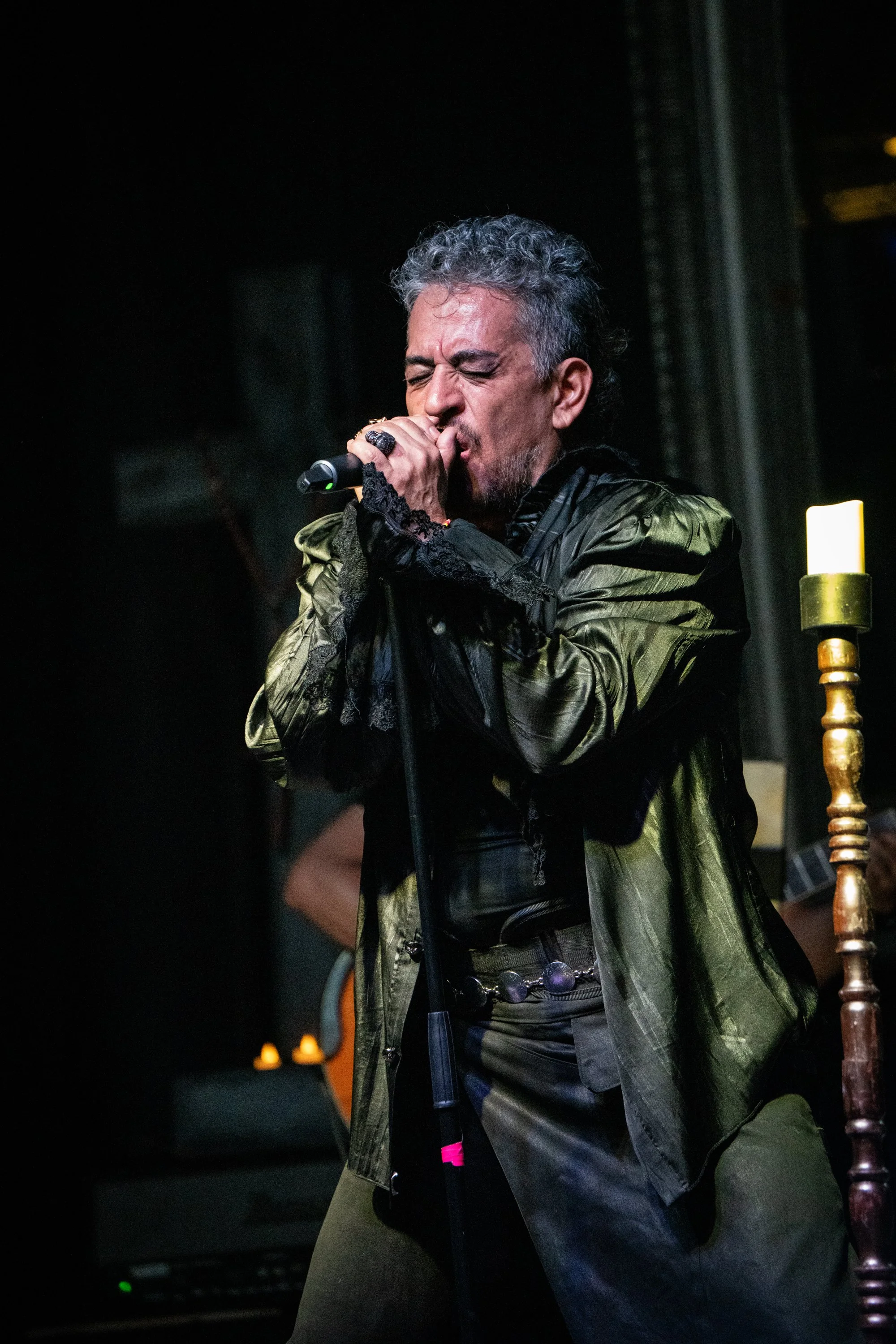 Un hombre cantando en un micrófono con fondo oscuro y candelabro dorado a la derecha.