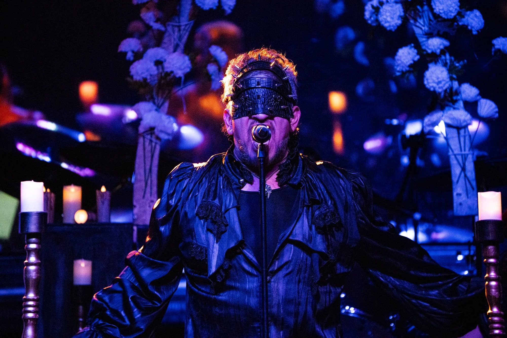 Un cantante con gafas negras y peinado elaborado se presenta en un escenario oscuro decorado con flores blancas y velas. Lleva una vestimenta negra y tiene un micrófono frente a la boca.