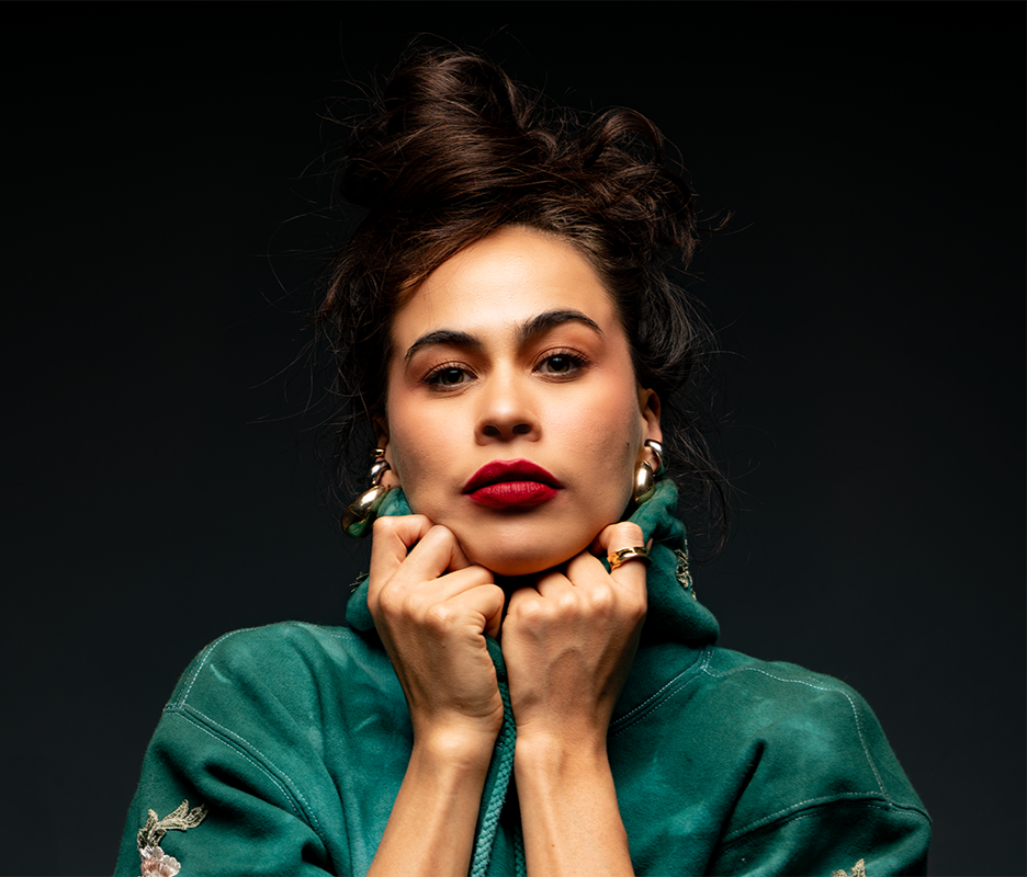 Retrato de una mujer con cabello oscuro y rizado, usando maquillaje llamativo, un suéter verde y accesorios plateados, posando con expresión segura en un fondo oscuro.