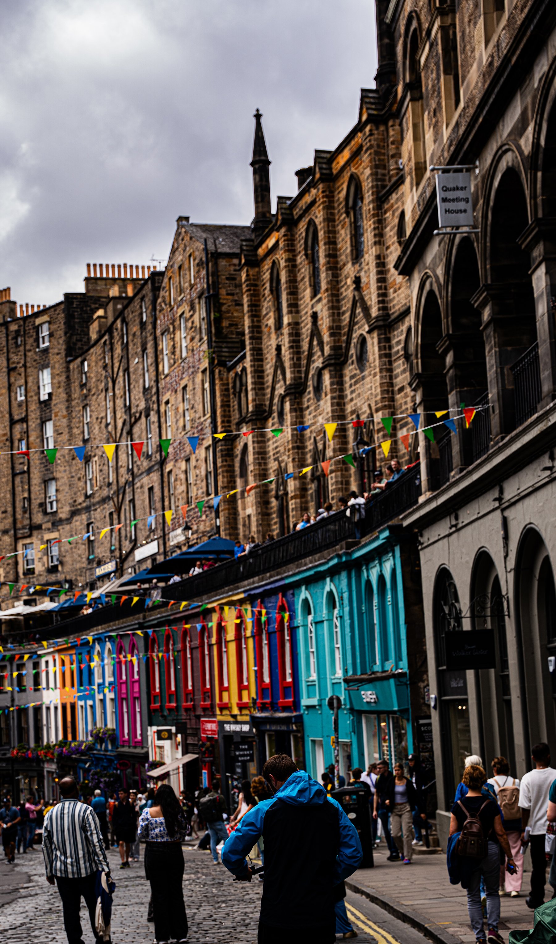 Vista de una calle concurrida con edificios antiguos en tonos marrón, decorada con banderas coloridas y con varias personas caminando por la acera, en un día nublado.