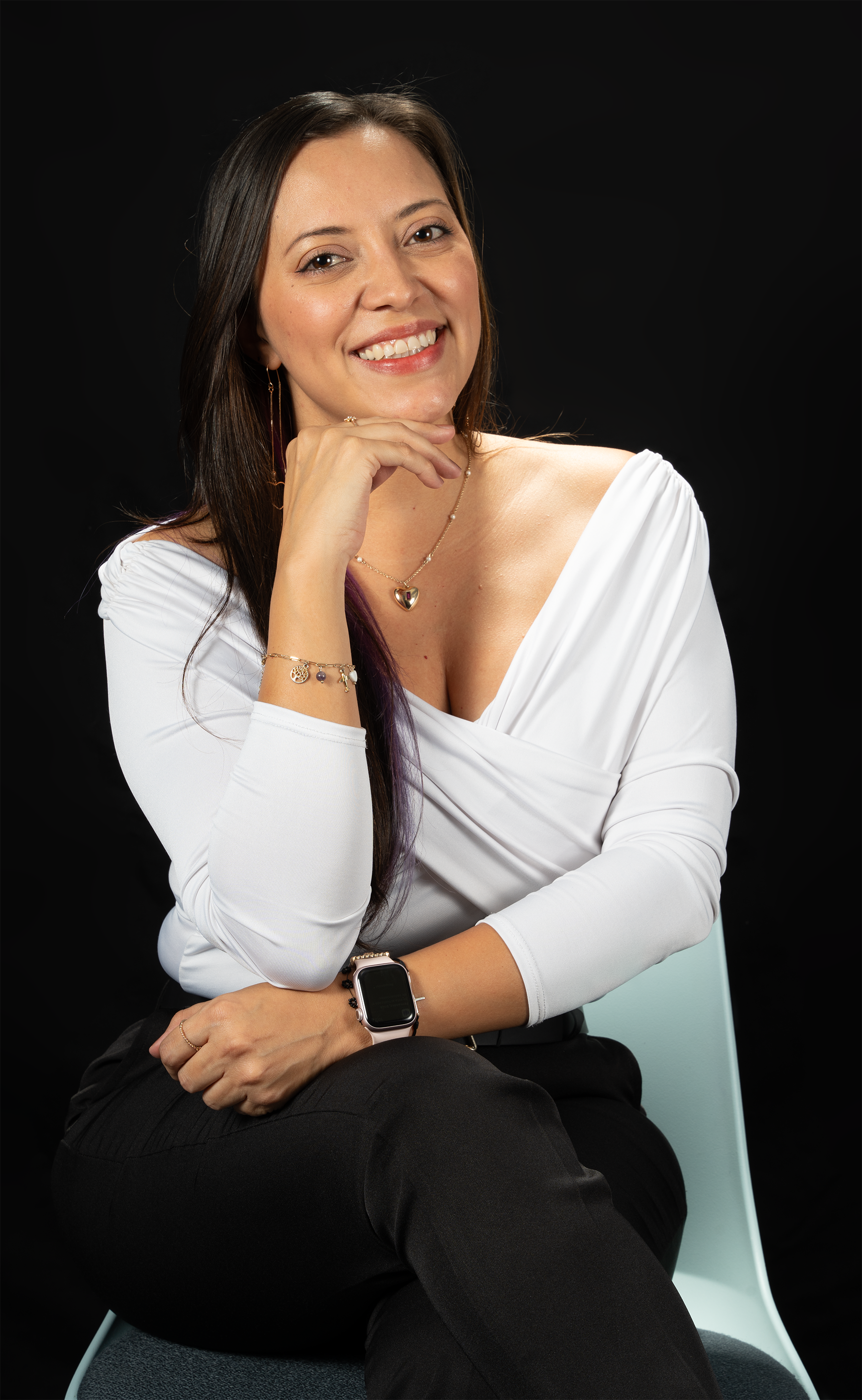 Una mujer con cabello largo y oscuro, sonriendo, lleva una blusa blanca con manga larga, joyas y un reloj inteligente, sentada en una silla, en fondo negro.