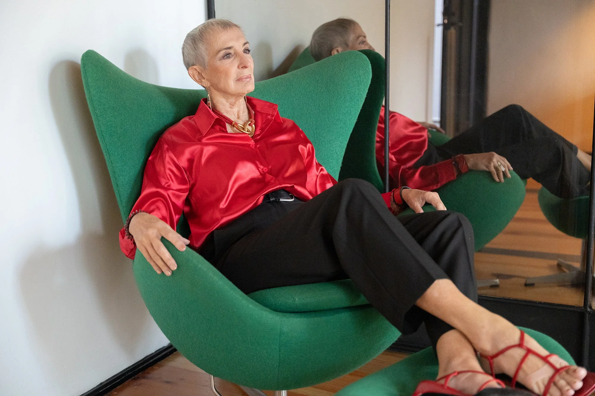 Mujer mayor con cabello canoso, vestida con blusa de satén roja, sentado en una silla verde moderna, con expresión serena y mirando hacia adelante en un ambiente interior.