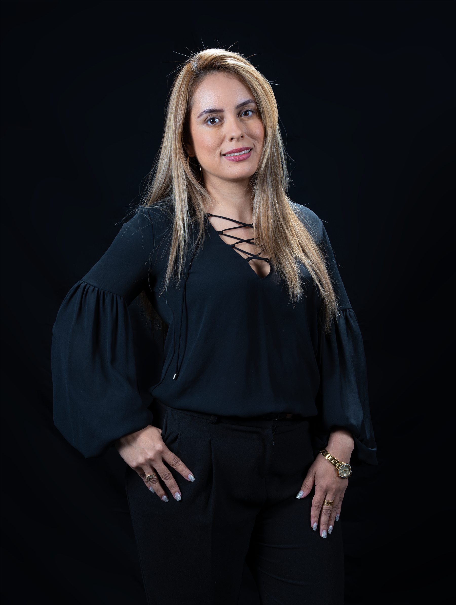 Retrato de una mujer con cabello rubio, ropa negra y fondo negro, con joyería en la mano y en la muñeca.
