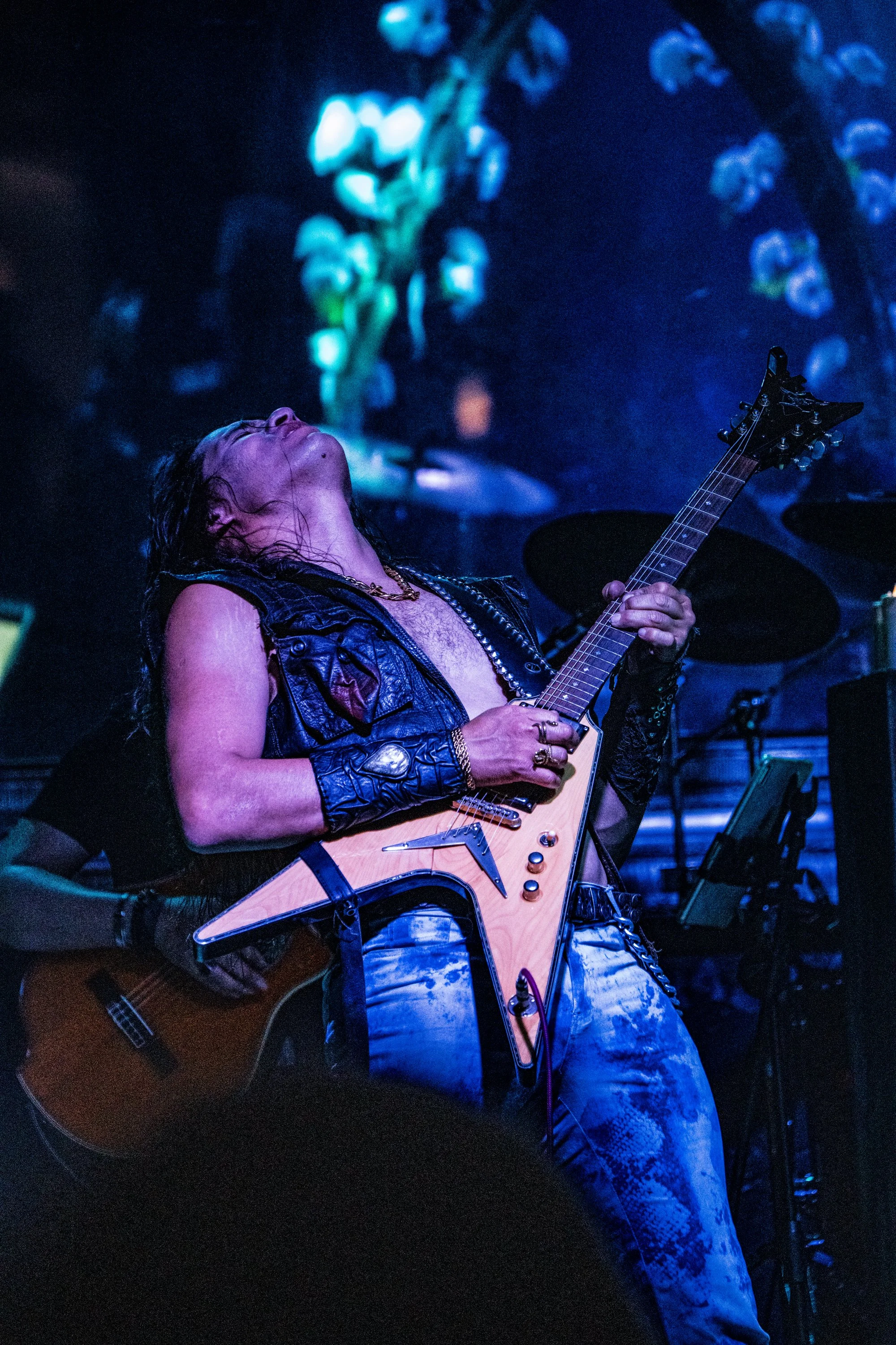 Un músico tocando una guitarra eléctrica de forma apasionada en un escenario oscuro con iluminación azul. Lleva una chaqueta de cuero y jeans rasgados, con cabello largo y húmedo.