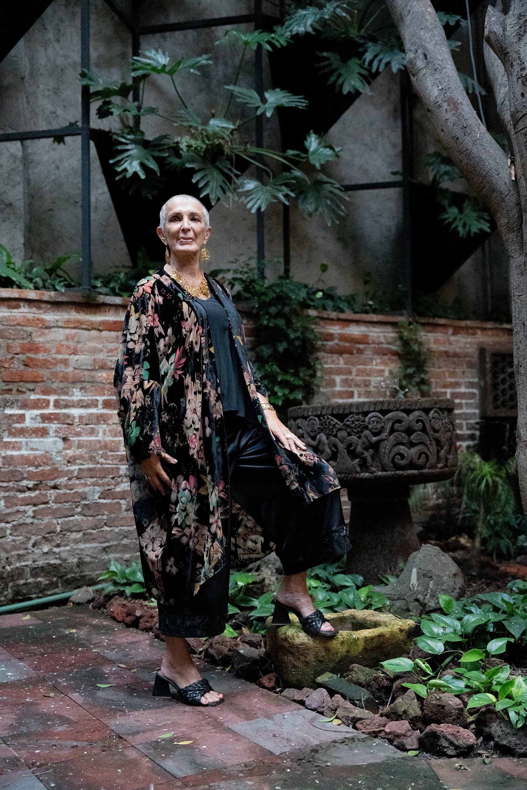 Mujer mayor con cabello castaño claro y vestido de flores negro con detalles dorados, de pie en un jardín, rodeada de plantas verdes y un muro de ladrillos, con árboles y una fuente decorativa.