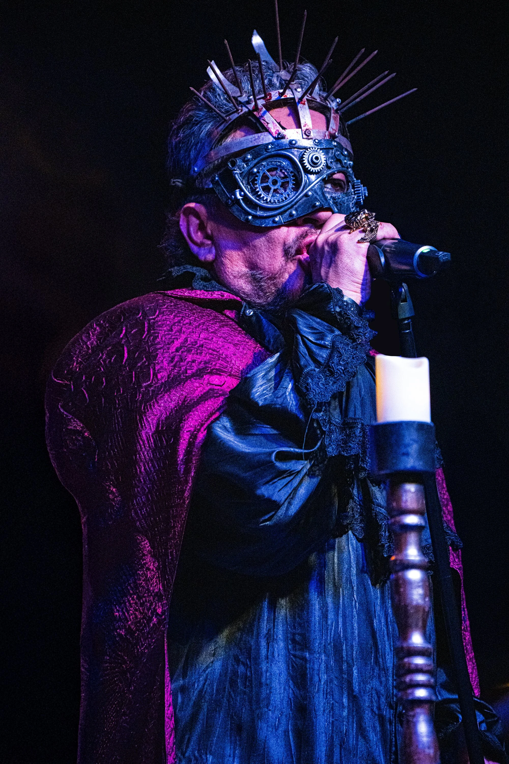 Hombre cantando con máscara de engranajes y cuchillas en la cabeza, vestido con ropa dorada y capa púrpura, en escenario oscuro.