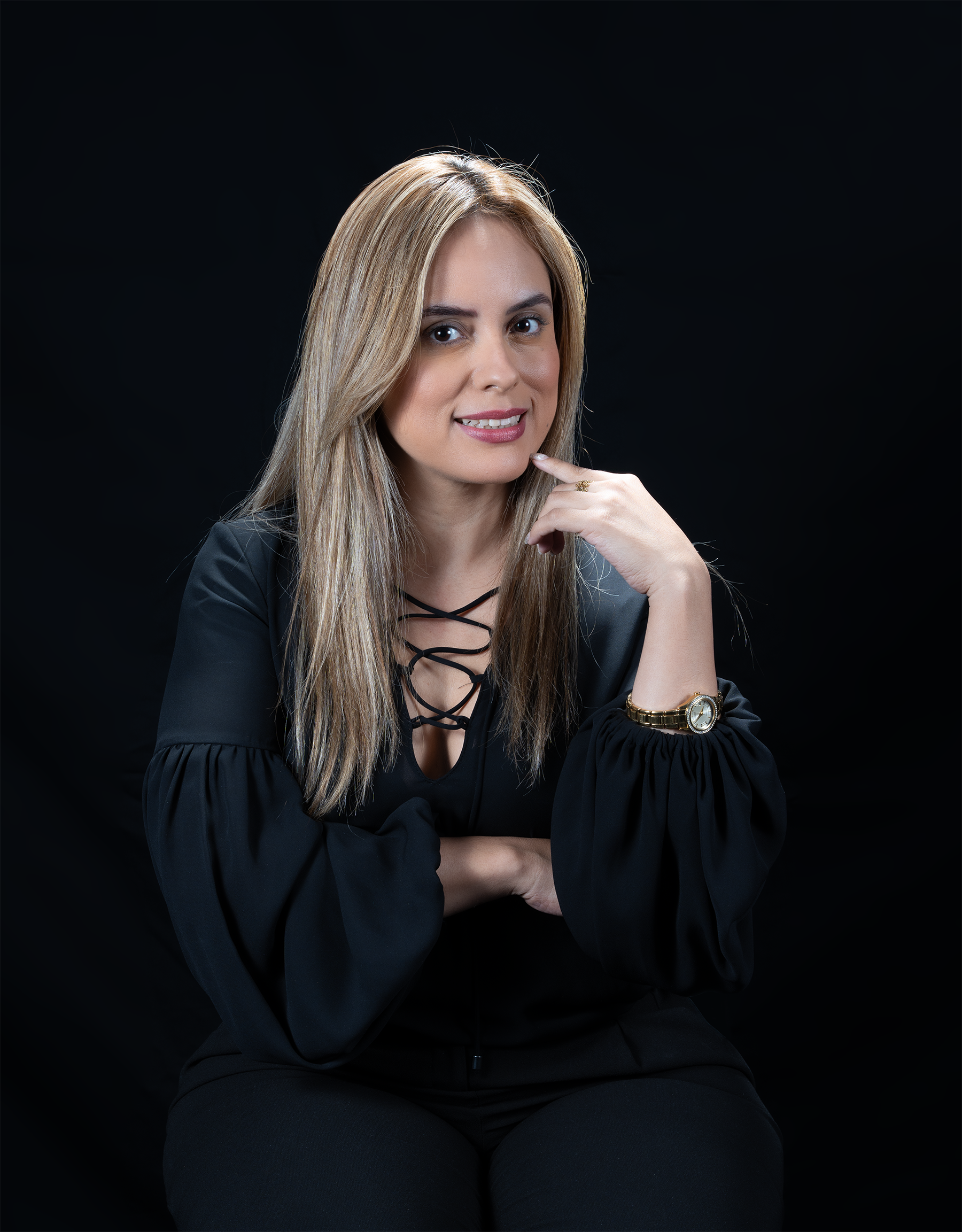 Mujer con cabello rubio y expresión pensativa, usando blusa negra con cordón en el escote, con reloj dorado en la muñeca izquierda, fondo negro.