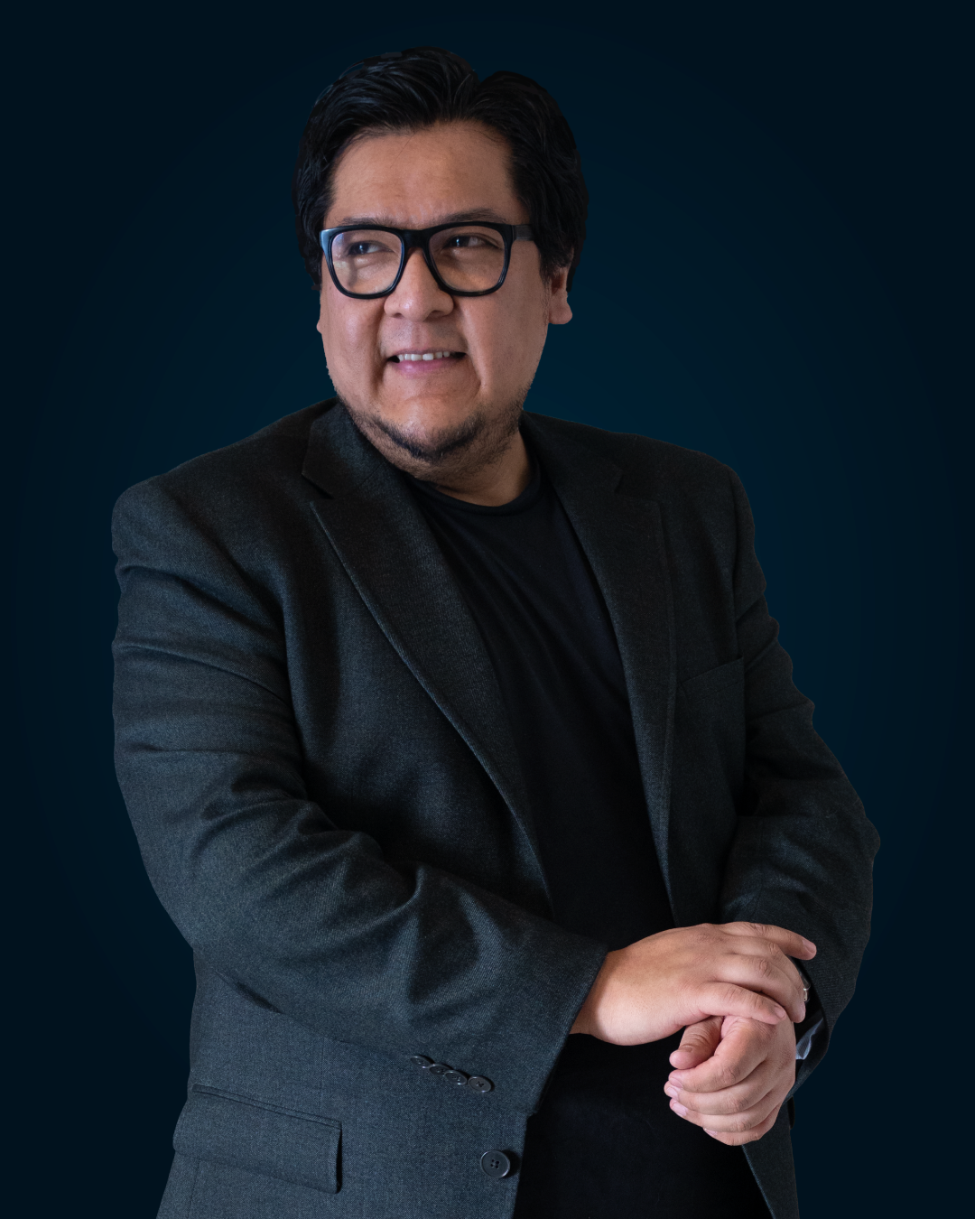 Hombre con gafas, cabello oscuro y barba, vestido con saco negro y camiseta negra, posando con las manos cruzadas y expresión pensativa, fondo azul oscuro.