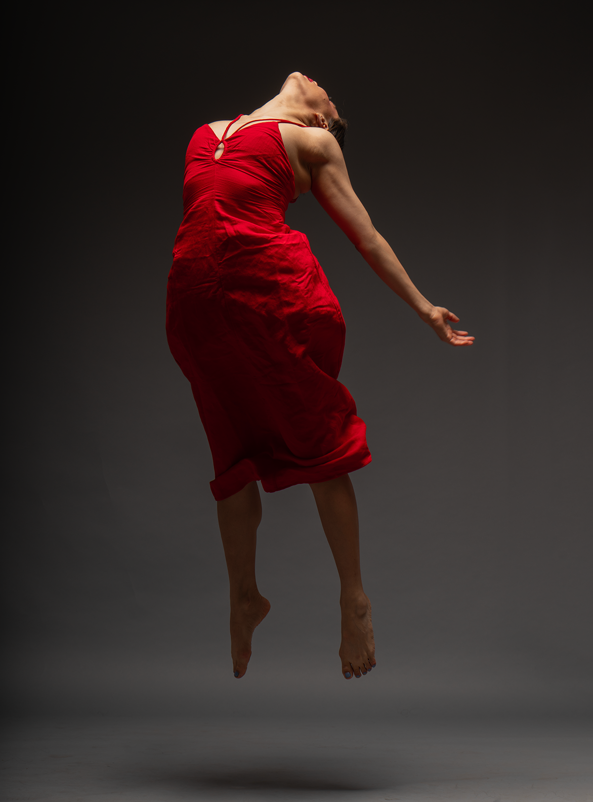 Mujer en vestido rojo en posición de danza, saltando con el cuerpo curvado hacia atrás y brazos extendidos, fondo gris oscuro.