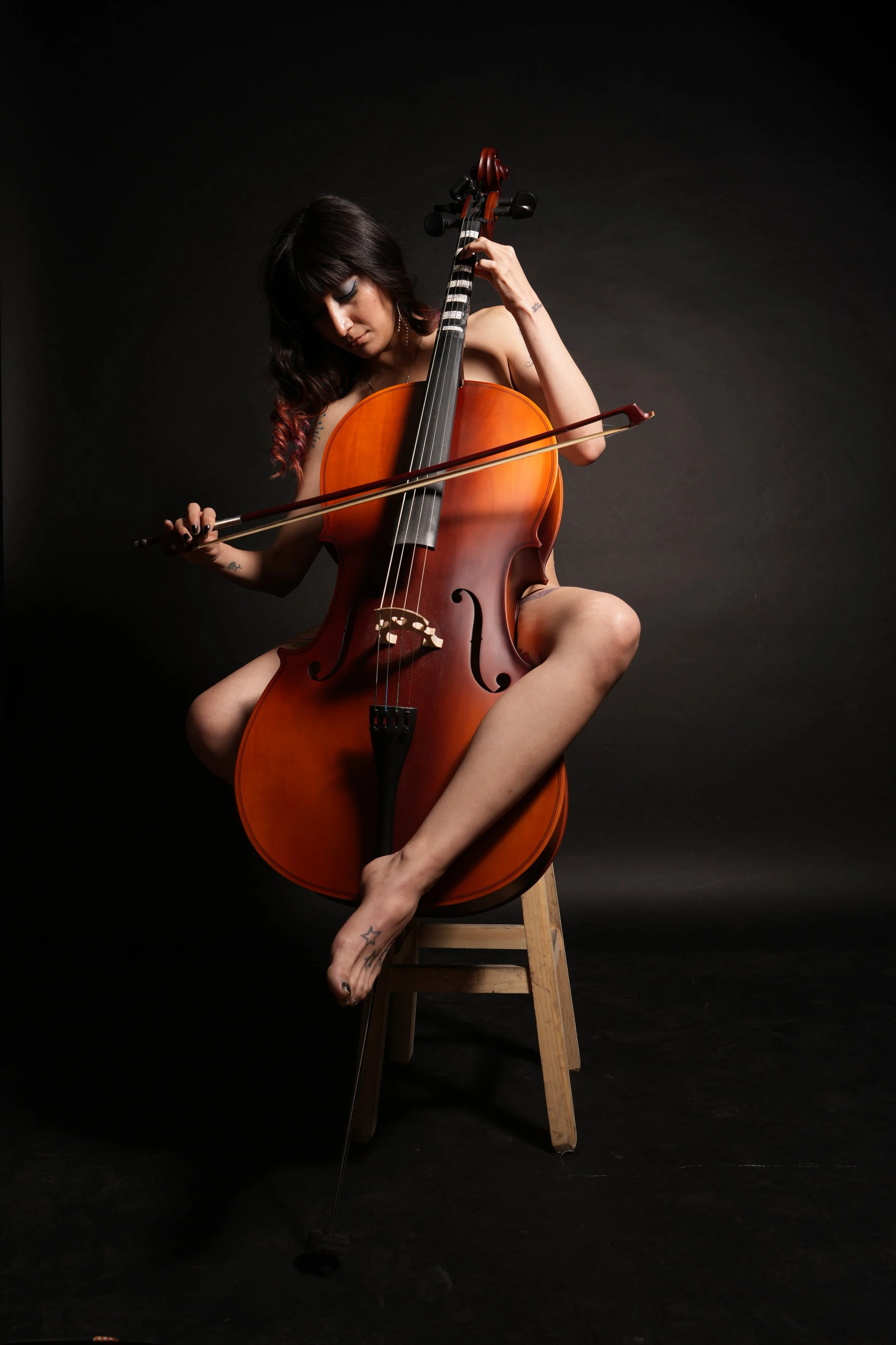 Mujer joven tocando un violonchelo en un fondo negro, sentada en una silla de madera, con cabello oscuro, maquillaje y tatuajes en sus brazos y pies.