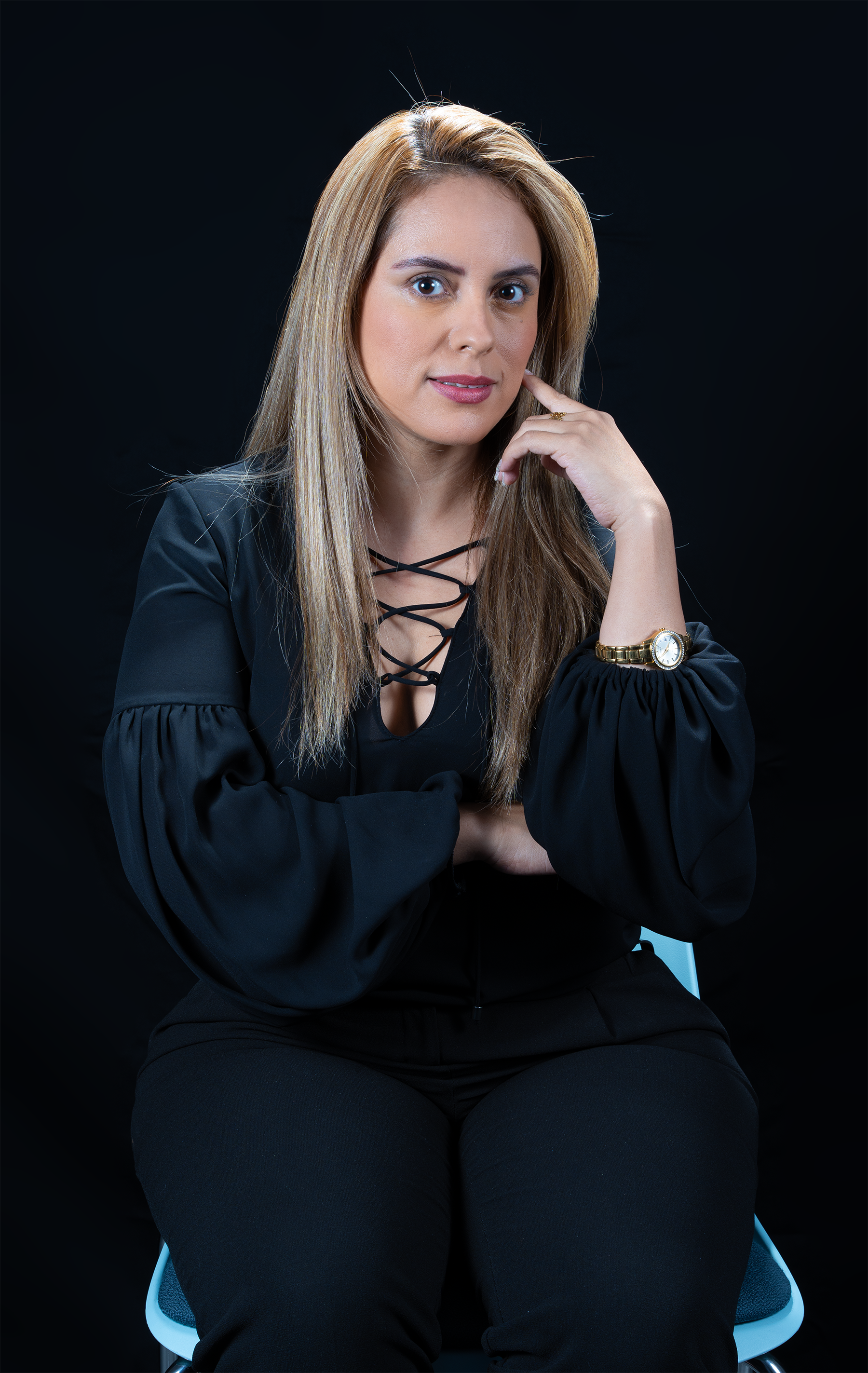Retrato de una mujer sentada en una silla, con cabello rubio y largo, vestida con blusa negra con cordones y pantalón negro, con fondo negro y una expresión pensativa, tocándose la mejilla con la mano derecha y usando un reloj dorado en la muñeca izq