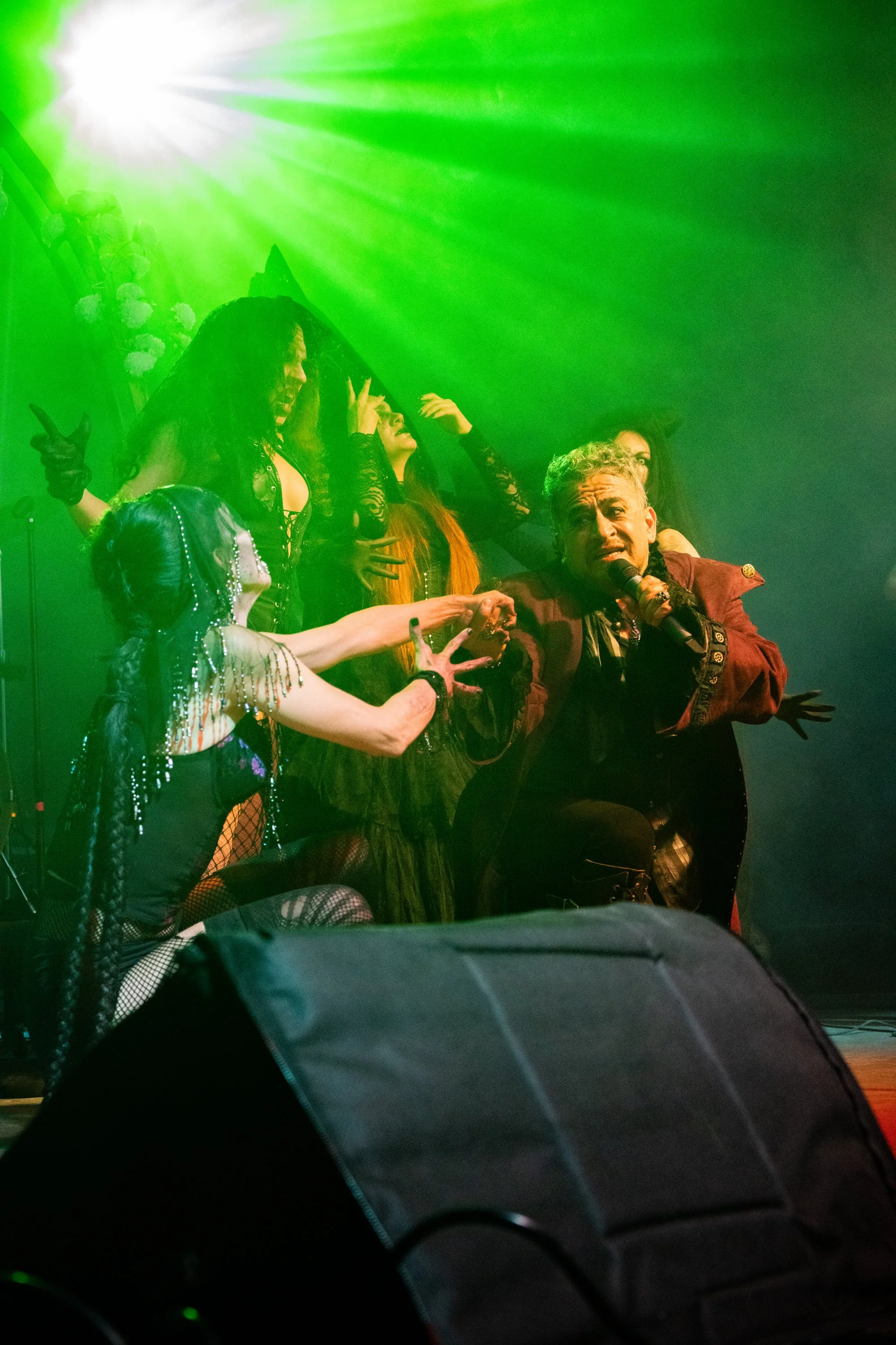 Grupo de personajes con vestimentas llamativas y maquillaje en un escenario con iluminación verde, uno de ellos sosteniendo un micrófono.