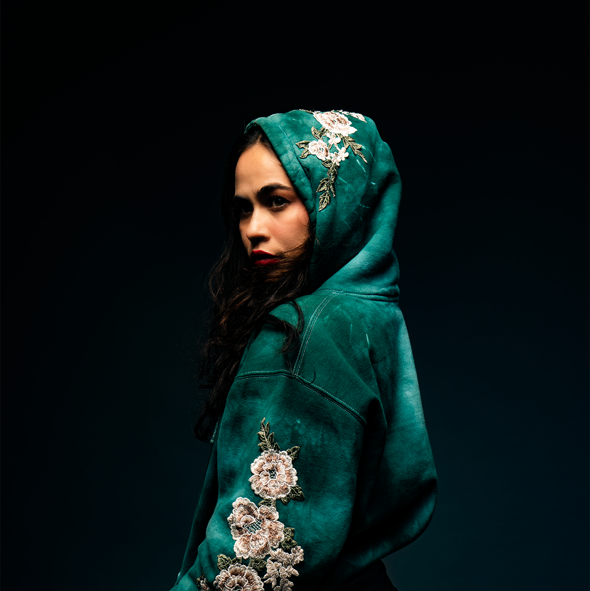 Mujer con hoodie verde con bordados florales oscuros en el fondo negro.