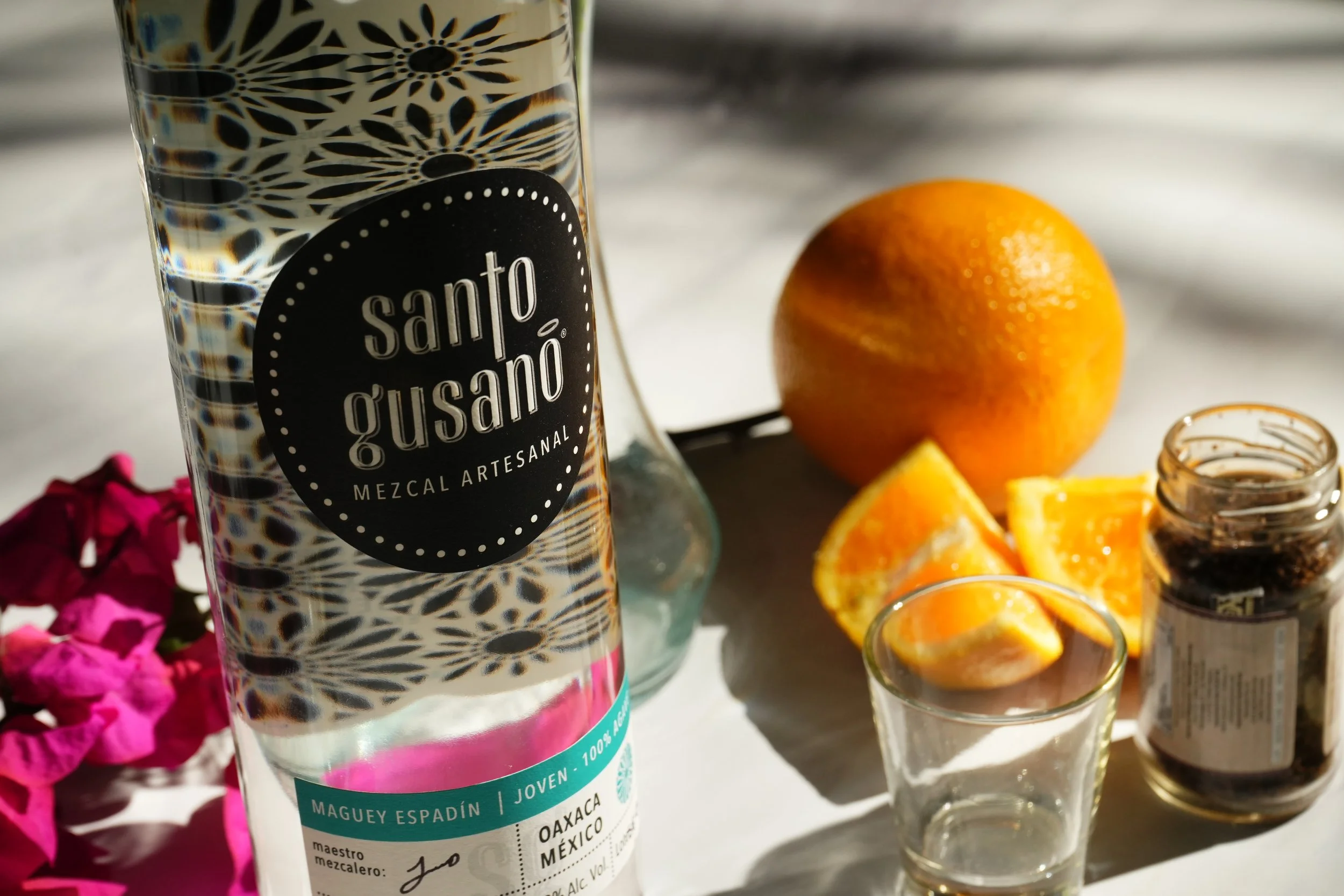 Botella de mezcal artesanal de Santa Gusano con etiqueta que indica que es de Oaxaca, México, junto a un vaso con jugo de naranja y una rodaja de naranja, y una pequeña botella con un líquido oscuro, rodeado de flores rosadas y pétalos.