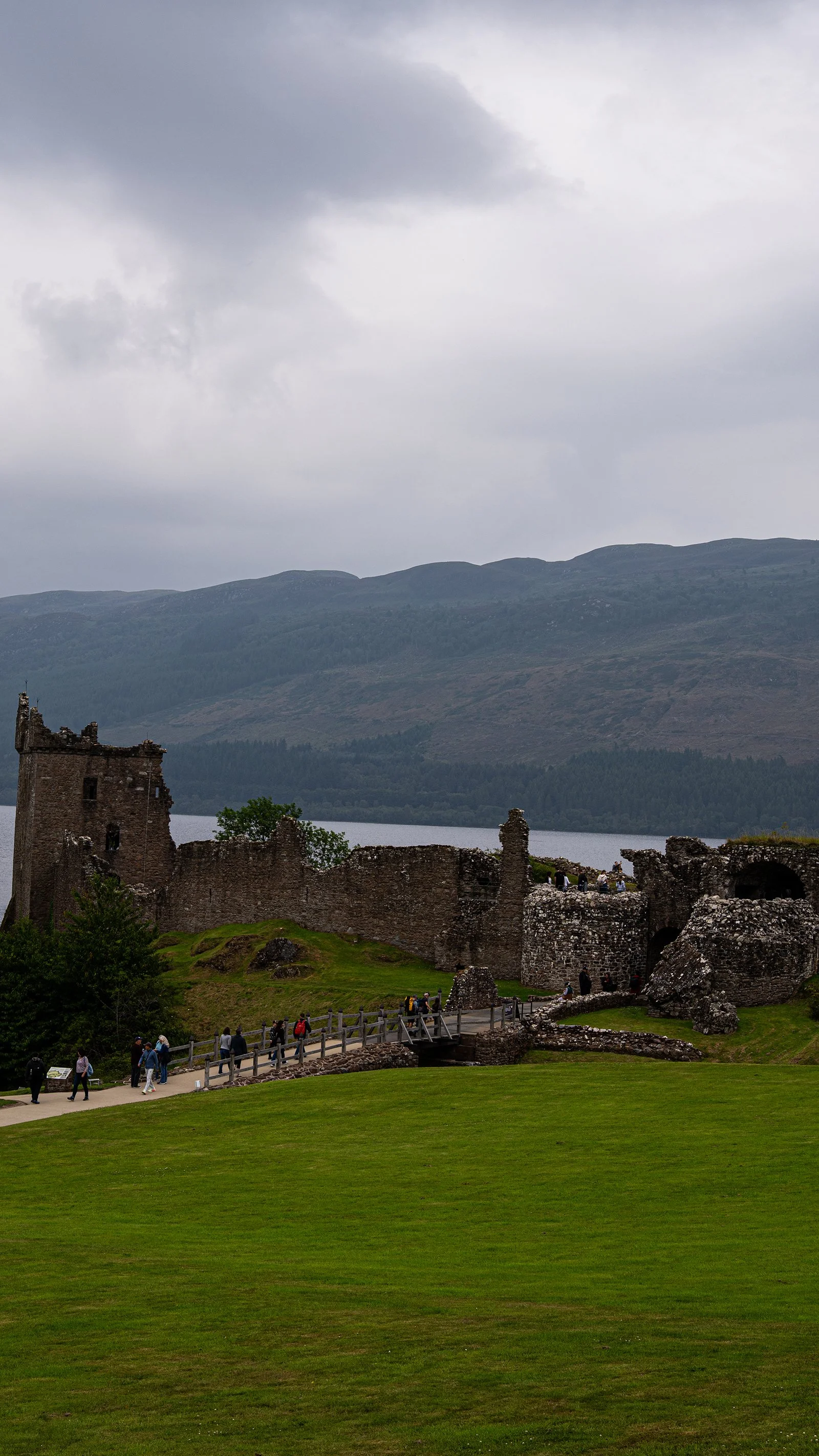 Ruinas del castillo de Eilean Donan con verdes praderas y montañas al fondo, bajo un cielo nublado.