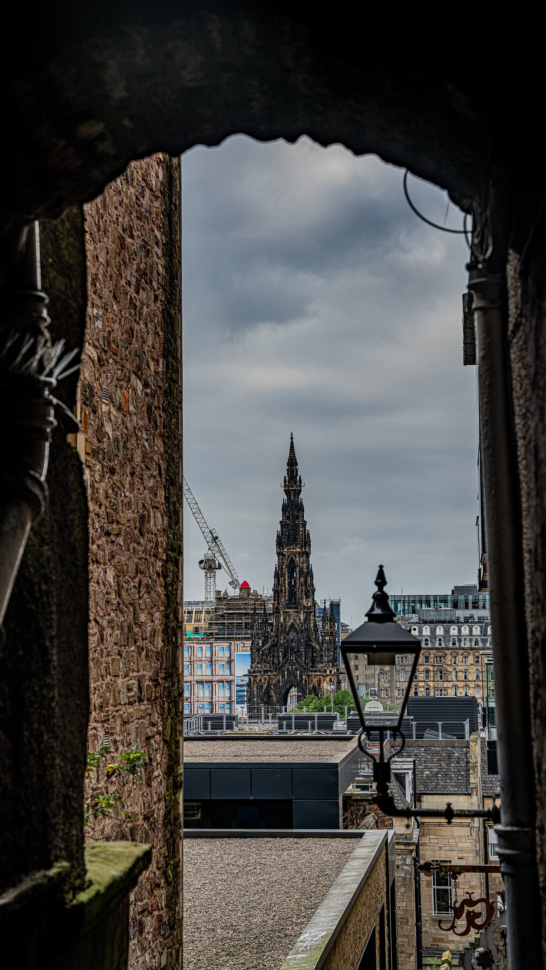 Vista de una calle en la ciudad con un edificio alto y una iglesia antigua al fondo, vista a través de un arco en una zona con edificios de piedra y un farol en primer plano. Nublado en el cielo.