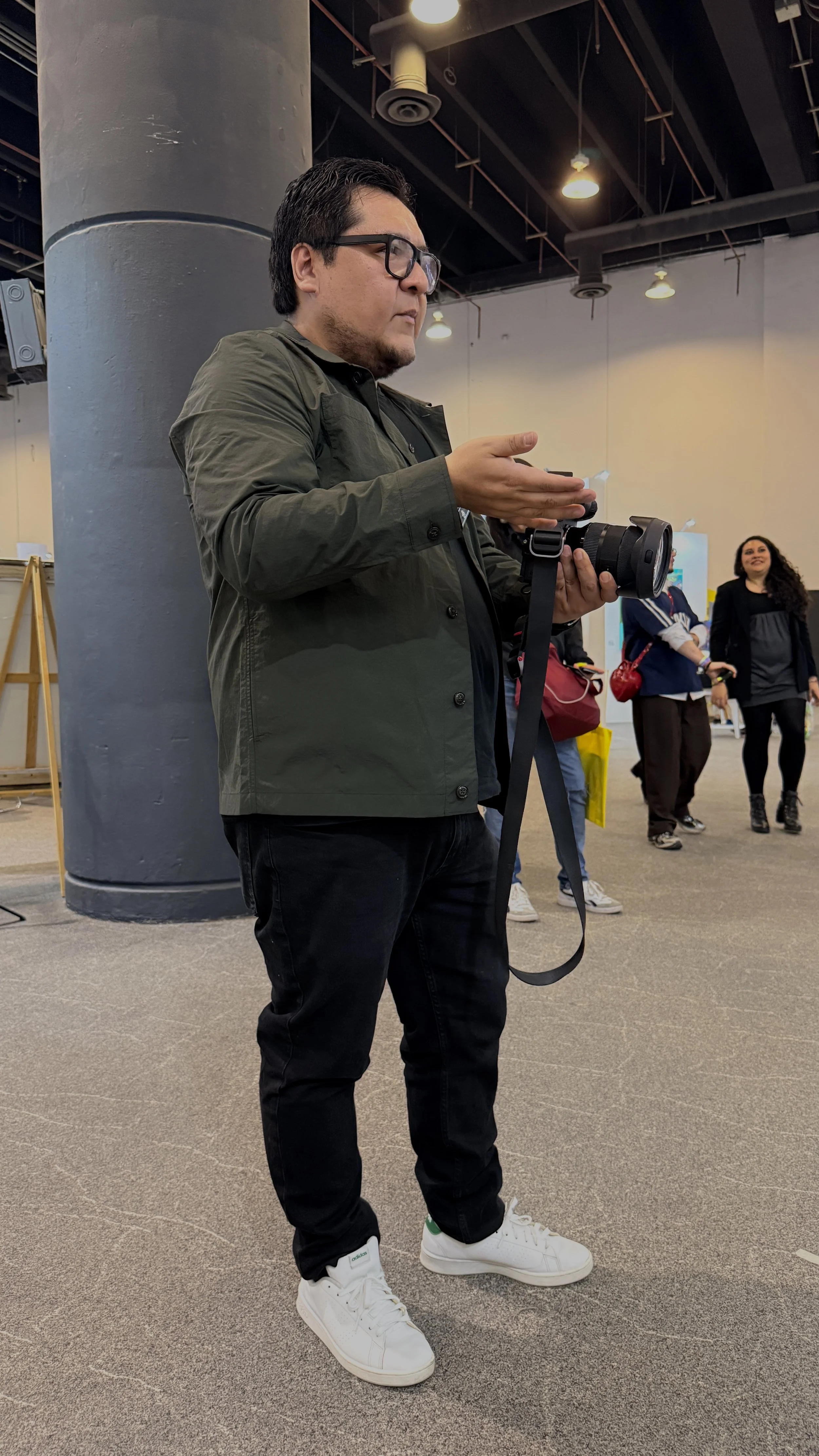 Hombre con gafas, chaqueta verde y pantalones oscuros sosteniendo una cámara en un lugar con grupo de personas al fondo.