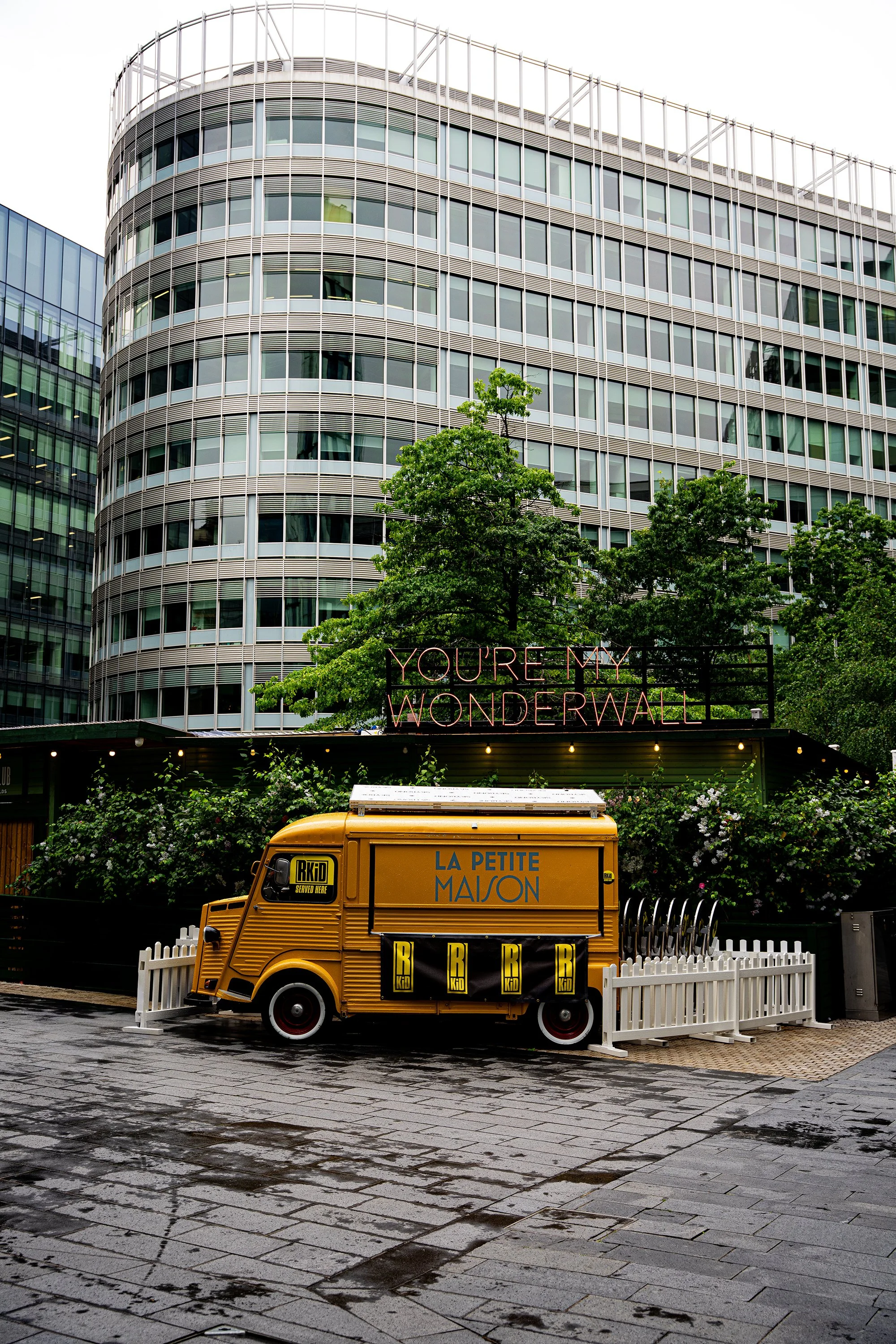 Edificio de oficinas de varias plantas con fachada de vidrio y estructura metálica. En la calle, un carrito amarillo de comida con cartel que dice "La Petite Maison" y cartel de BKR. En el fondo, cartel de neón que dice "You're my wonderwall". Hay ár