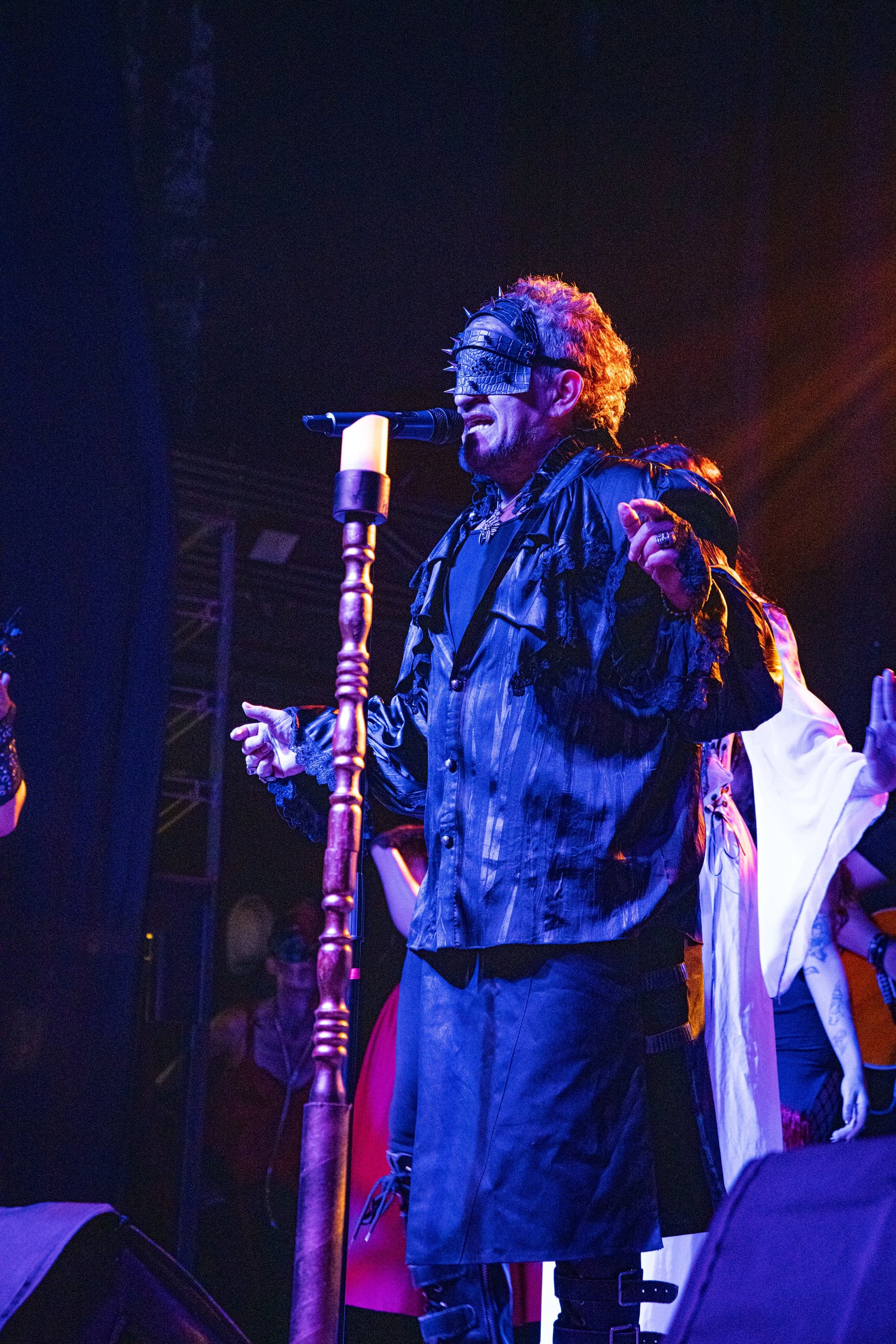 Hombre en escenario con máscara de cuero con púas, sosteniendo un micrófono y cantando con luces de colores, vestimenta oscura y accesorios metálicos.