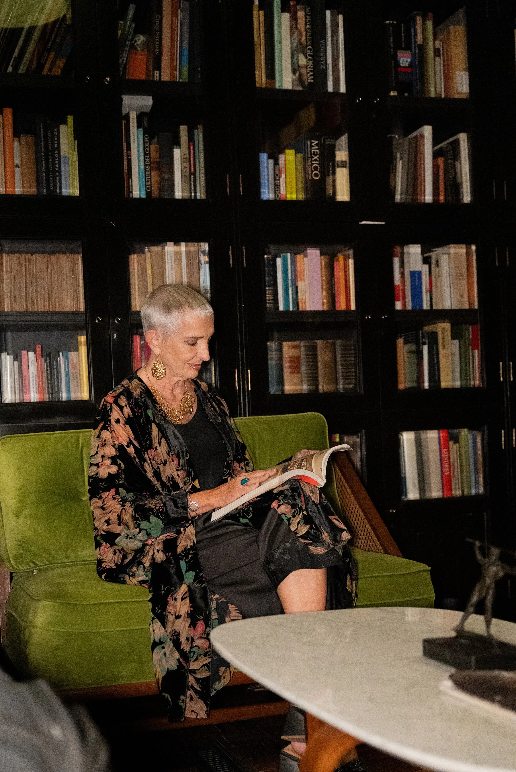 Una mujer de cabello corto y canoso, vestida con un vestido negro y un kimono con estampado floral, está sentada en un sillón verde, leyendo un libro en un espacio con estanterías llenas de libros en el fondo.