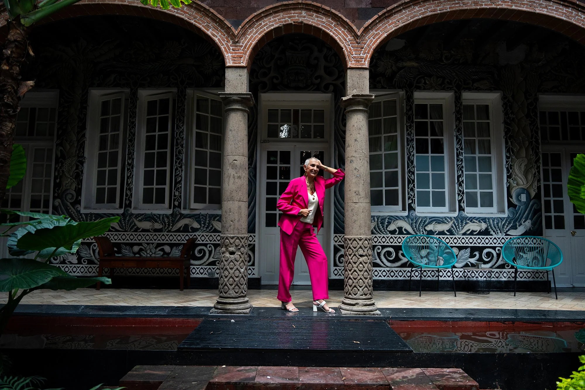 Una mujer sonriendo y luciendo un conjunto rosa brillante en un espacio con columnas, ventanas blancas y decoración con motivos de animales y naturaleza en las paredes.