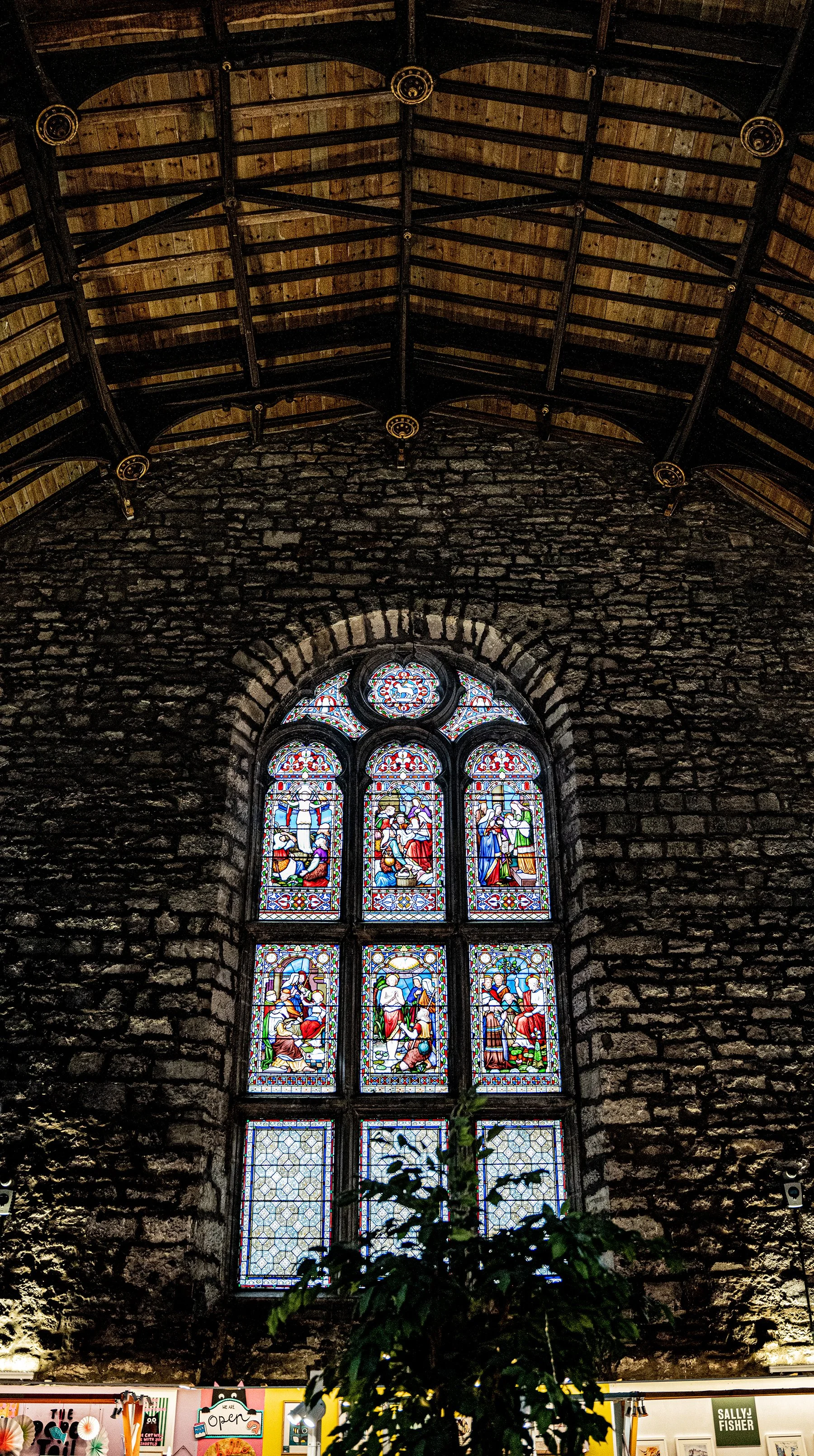 Ventana de vitrales en una pared de piedra dentro de una iglesia, con un árbol en primer plano y un cielo visible a través del vitral.