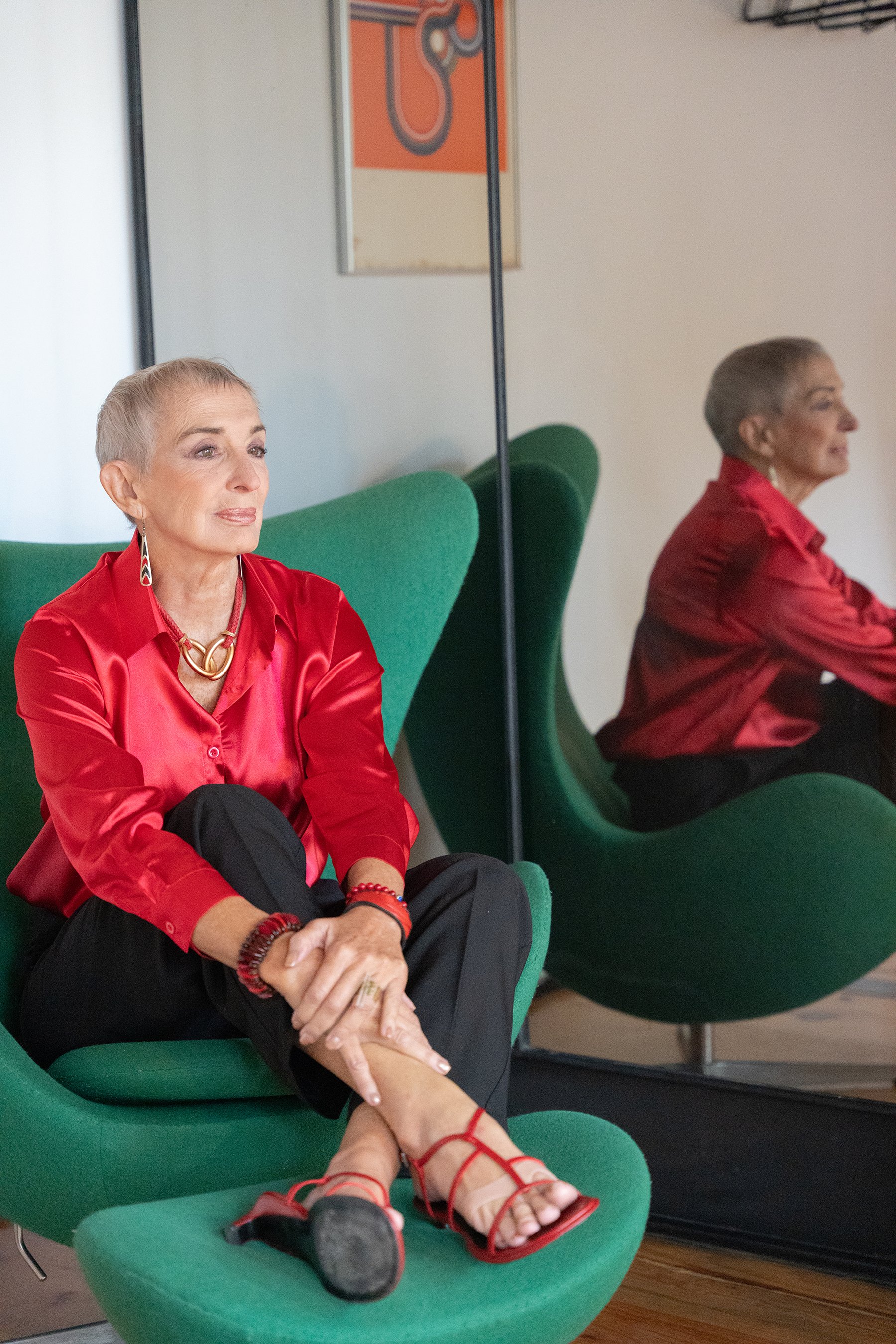 Mujer de cabello corto y canoso con ropa roja, gafas y accesorios, sentada en una silla verde, con reflejo en un espejo.