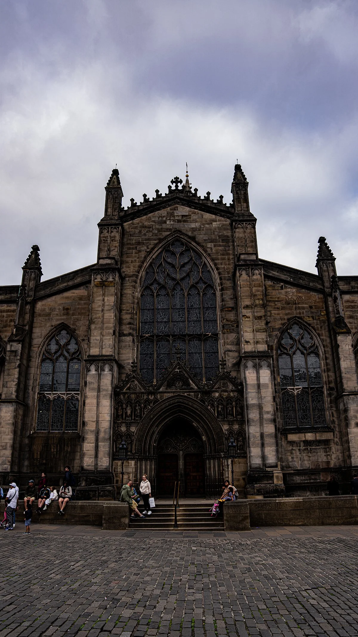 Fachada de una iglesia gótica con vitrales grandes, en piedra, con personas sentadas en los peldaños y caminando en la plaza frente a ella.