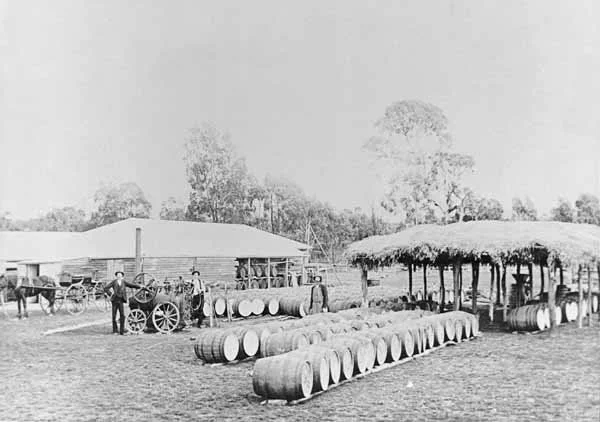 Old black and white photo of a historic rural scene with barrels, a horse-drawn wagon, and people standing near a thatched shelter.