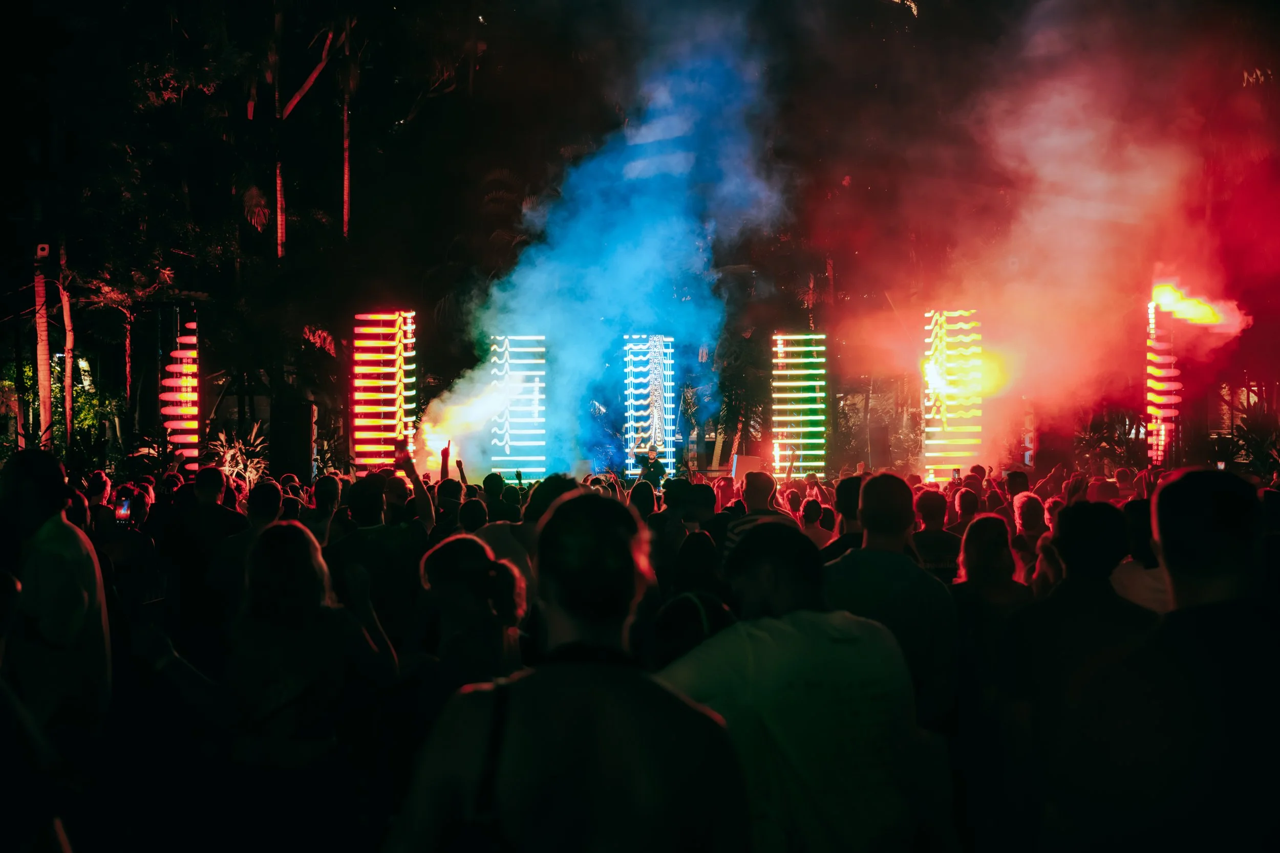 Crowd at a nighttime outdoor concert with colorful stage lights, smoke effects, and palm trees