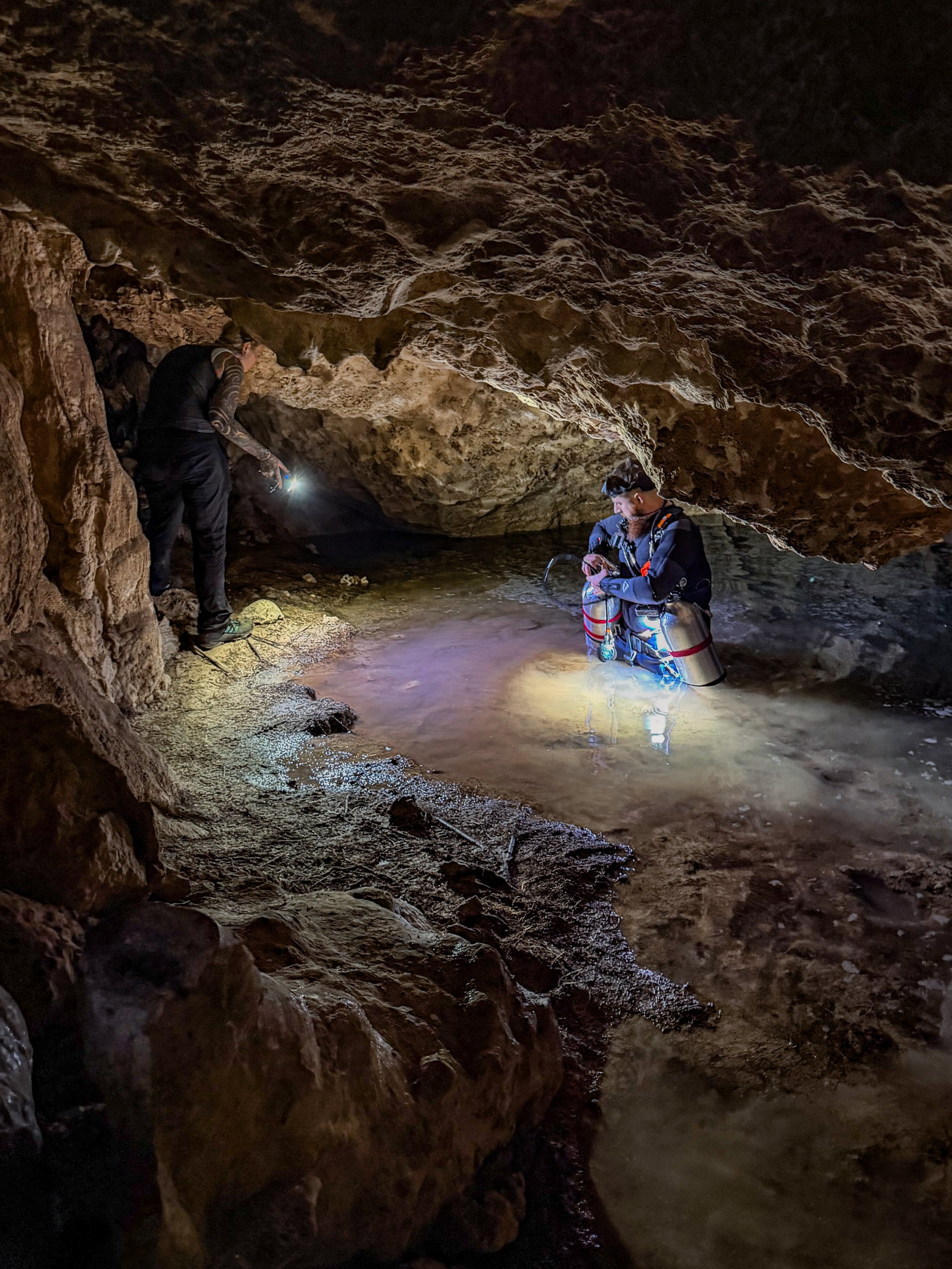 Two people exploring a dark cave with water, they are both looking into the distance, and the cave has rough, brown rock formations.