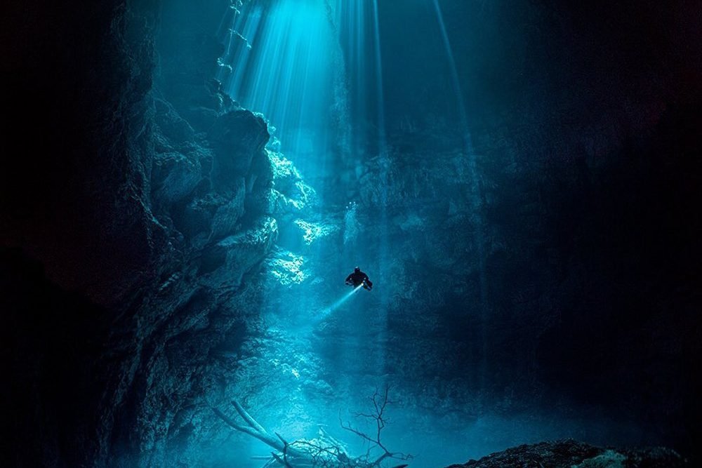 An underwater scene of a scuba diver exploring a deep cavern with sunlight streaming through the opening above.