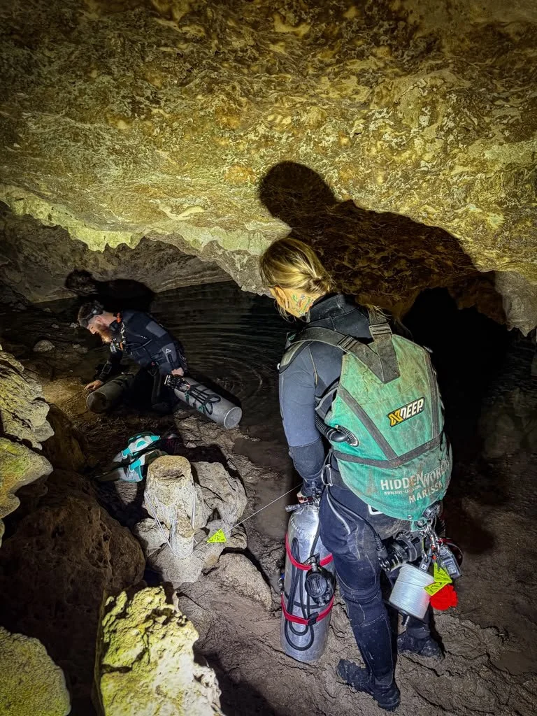 Two explorers in caving gear exploring a narrow underground cave passage with rocky walls, equipped with scuba gear, flashlights, and ropes.