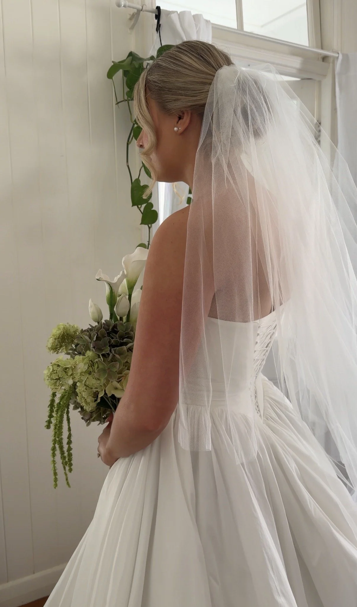 Side view of a bride in a white wedding dress with a tulle veil, holding a bouquet of white and green flowers, standing indoors near a window with white wood paneled walls.
