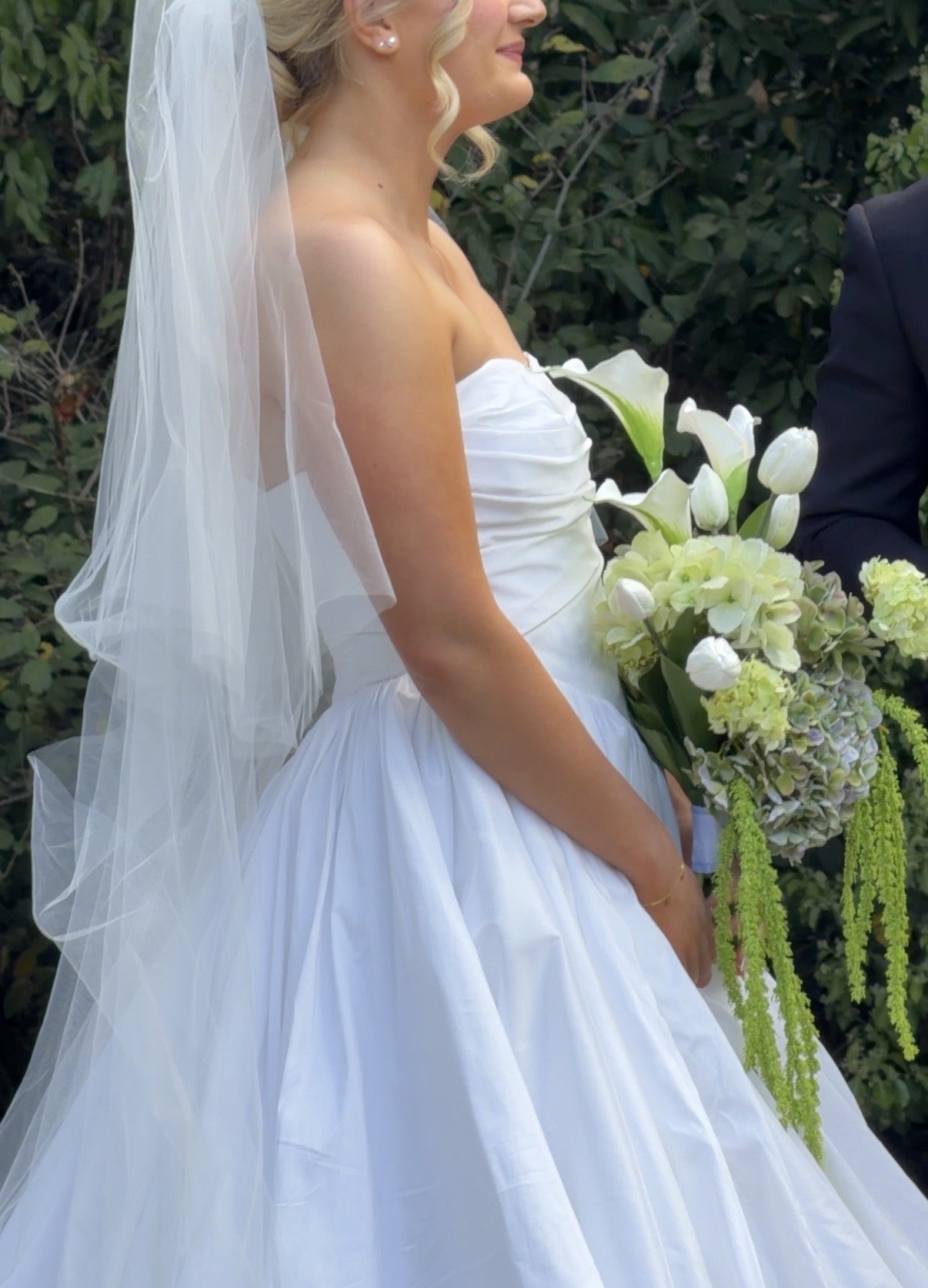 A bride in a white wedding dress holding a bouquet of white and green flowers, standing outdoors with green foliage in the background.