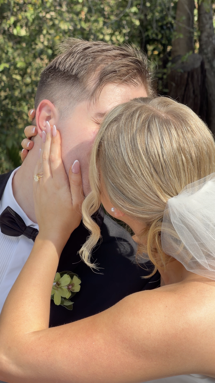 A newlywed couple sharing a kiss outdoors, with the bride's hand gently holding the groom's face.