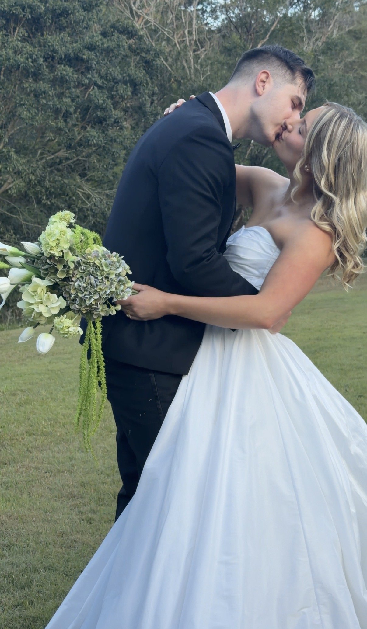 A bride and groom kiss outdoors during their wedding, with the bride holding a bouquet of green and white flowers.