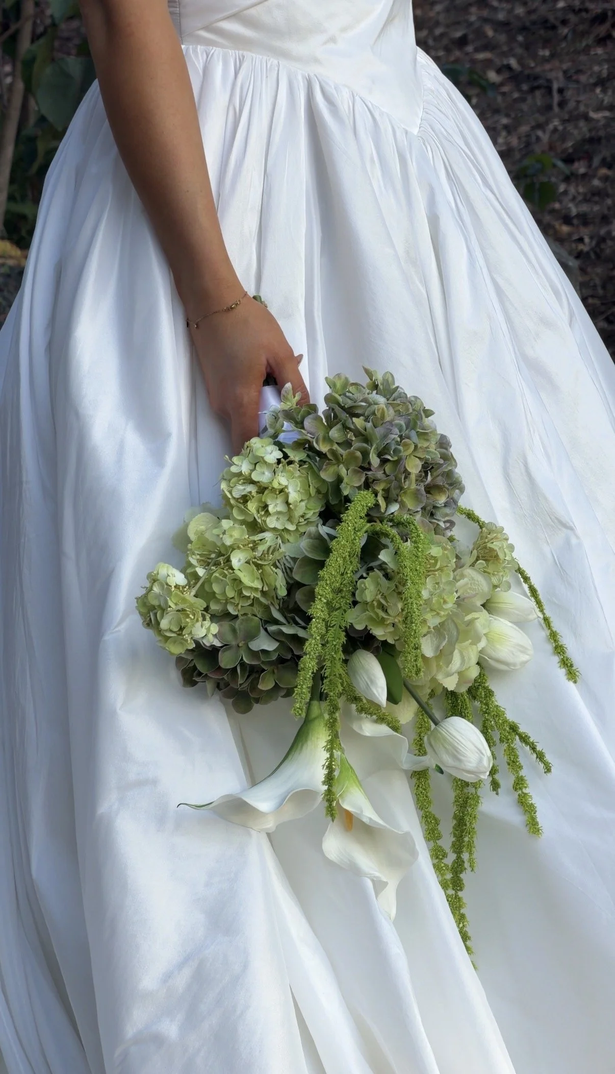 A woman in a white wedding dress holding a bouquet of green and white flowers, including calla lilies and hydrangeas, outdoors.