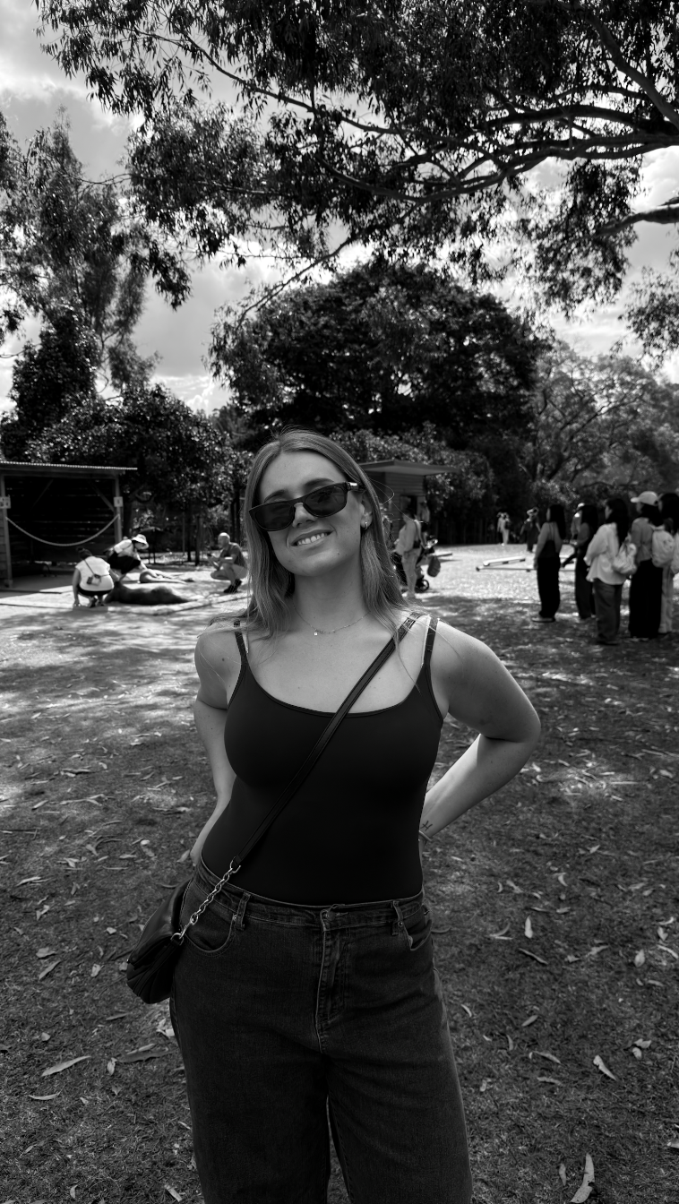A woman with long hair wearing sunglasses, a black tank top, and jeans, smiling at the camera in a park with trees and other people in the background.