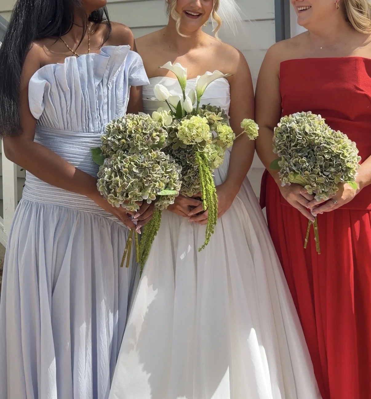 Three women in colorful dresses holding bouquets of flowers, one in white, one in blue, and one in red, standing outdoors.