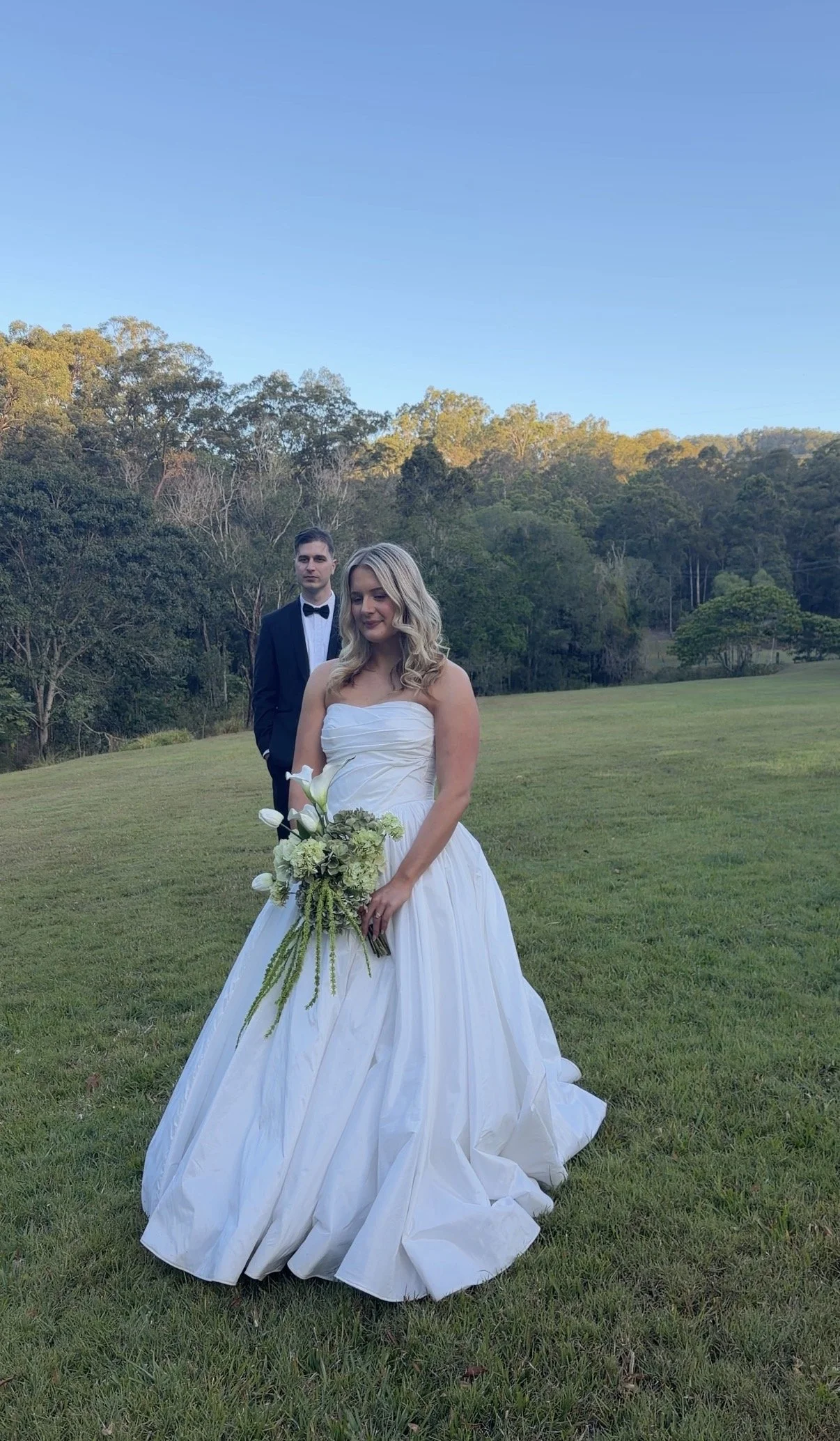 A bride in a white wedding gown holding a bouquet of white and green flowers, standing on a grassy field with a forested hill and blue sky in the background. A man in a dark suit and bow tie stands behind her.