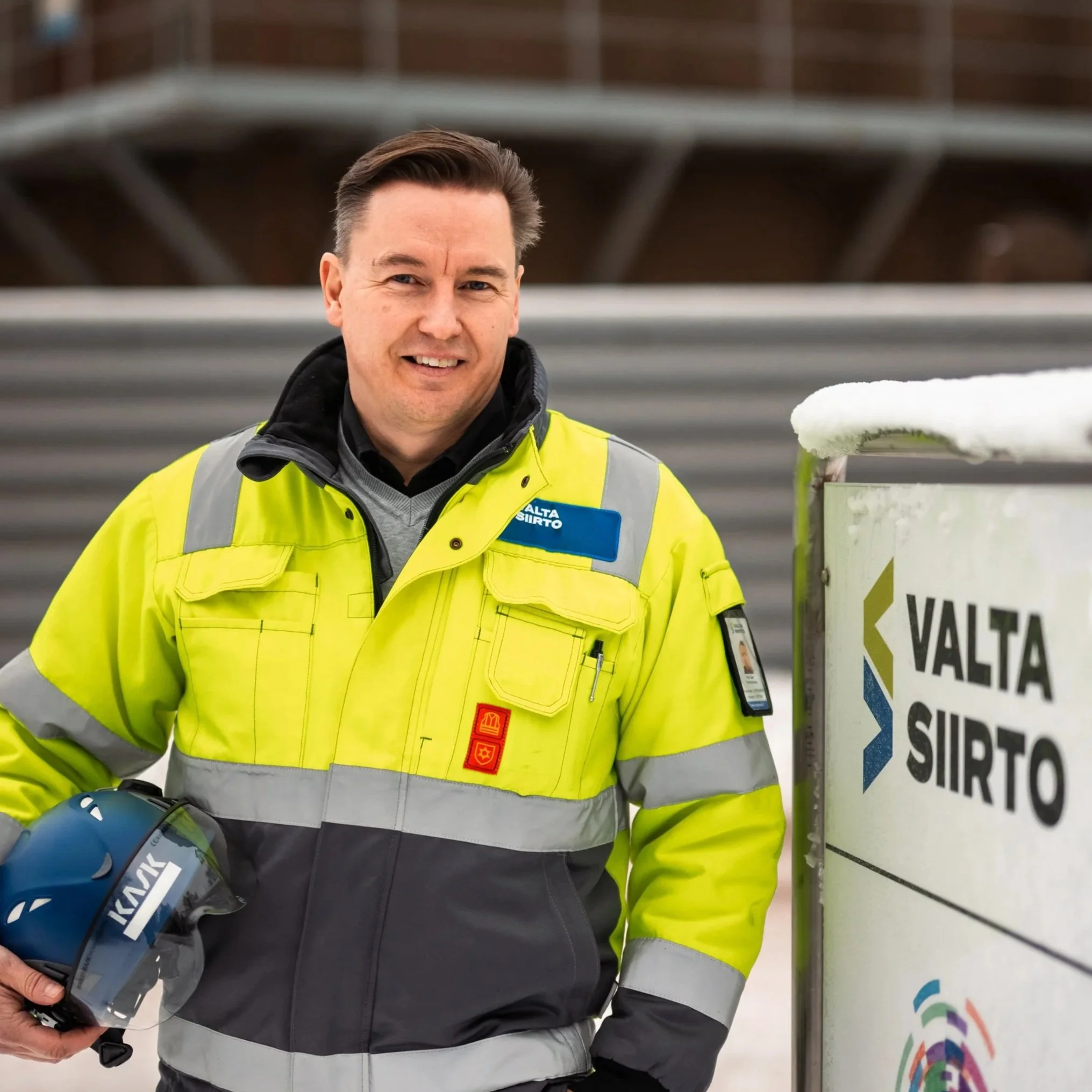 Mieshenkilö, joka on pukeutunut kelta-vihreään työvaatteeseen, seisoo ulkona ja pitää kypärää kädessään.