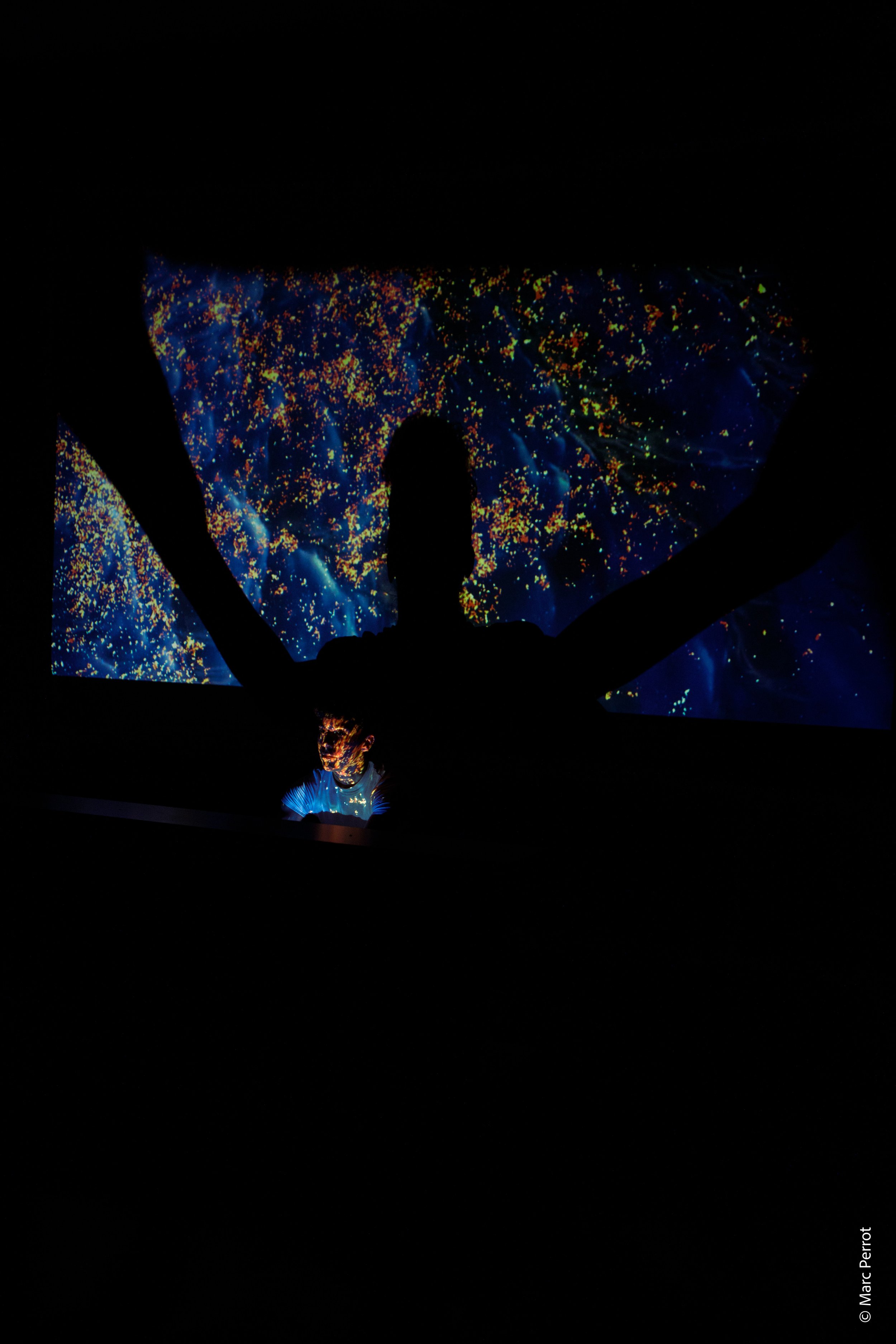 Une installation artistique avec un écran montrant un fond bleu et des points colorés en jaune, rouge, et orange. Un mannequin, éclairé par la lumière de l'écran, porte une robe bleue illuminée, avec un fond noir.